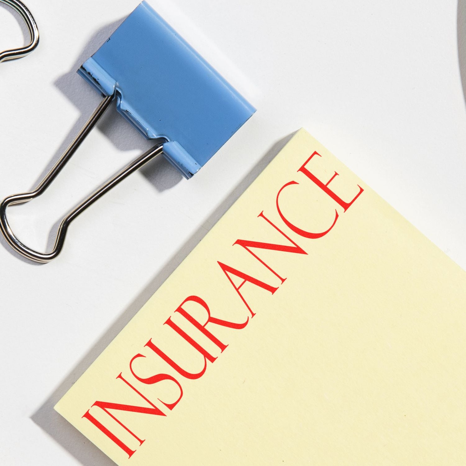 A blue binder clip and a yellow paper stamped with INSURANCE in red using the Insurance Rubber Stamp, placed on a white surface.