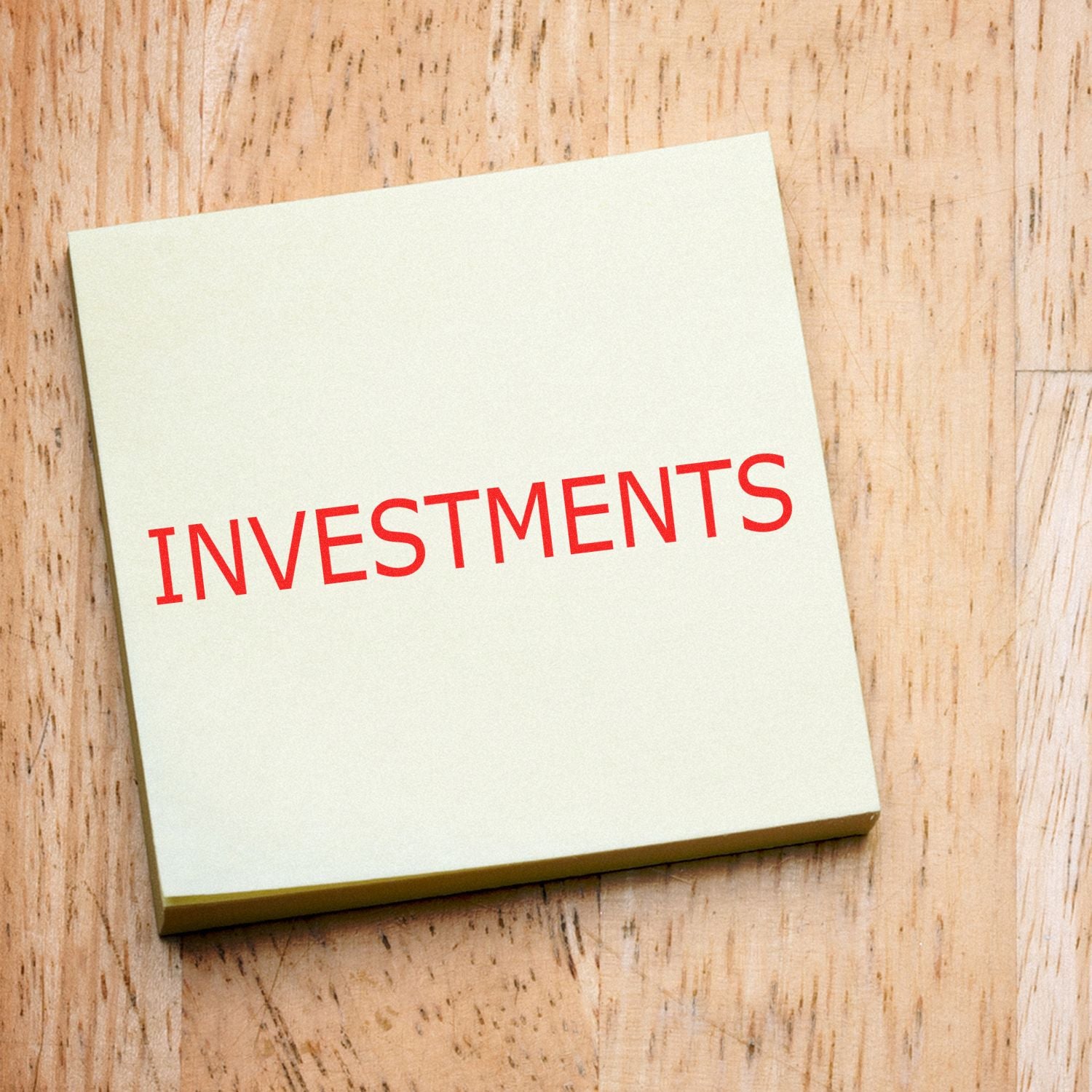 Investments Rubber Stamp in red ink on a yellow sticky note, placed on a wooden surface.