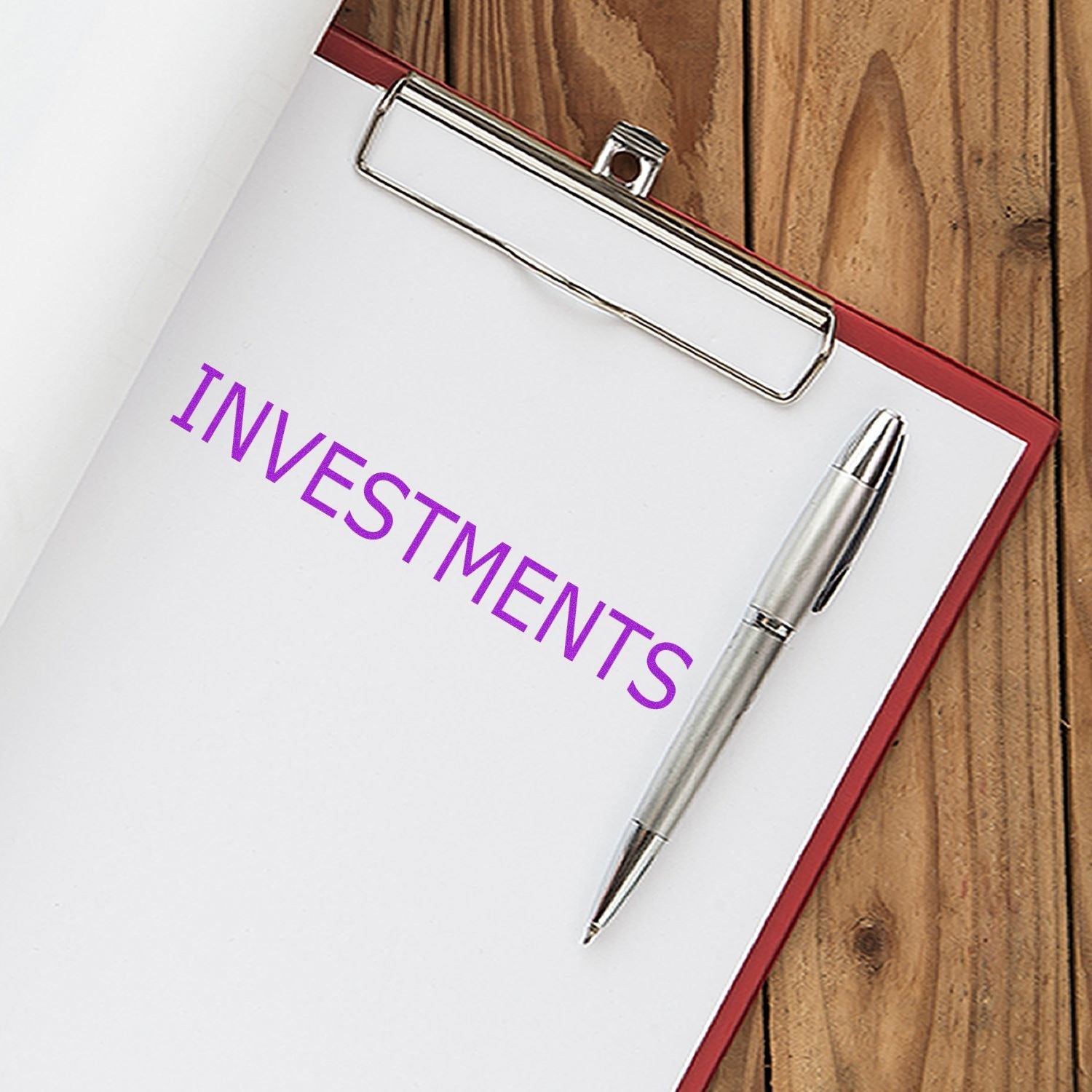 Clipboard with paper stamped INVESTMENTS using Self Inking Investments Stamp, next to a silver pen on a wooden surface.