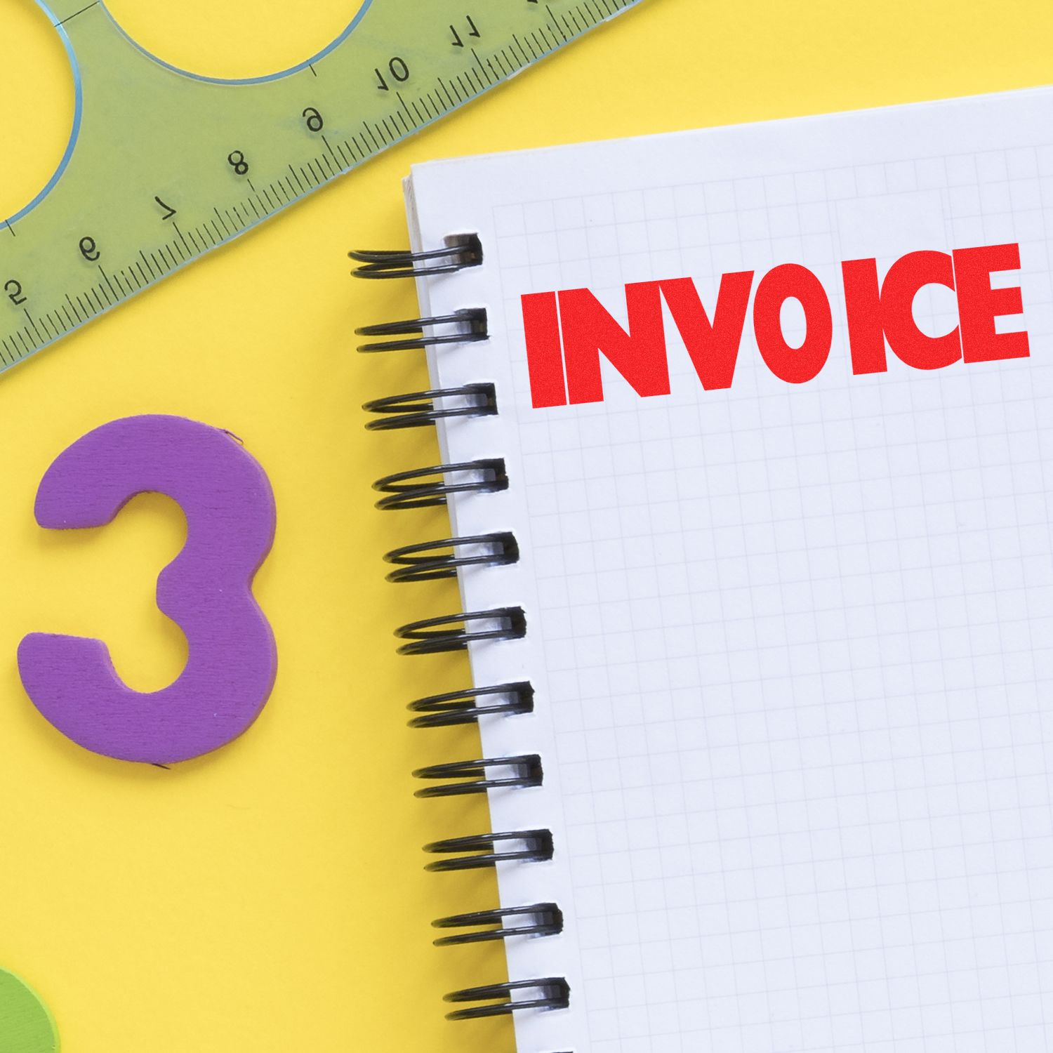 Large Self Inking Invoice Stamp used on a spiral notebook with INVOICE in red, next to a ruler and a purple number 3 on a yellow background.