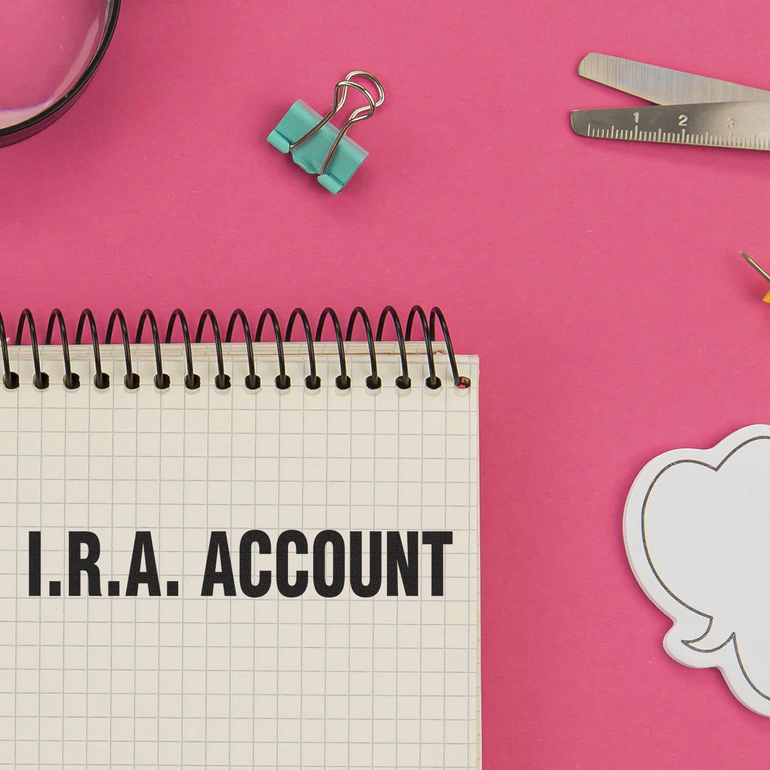 Large I.R.A. Account Rubber Stamp used on a notebook with I.R.A. ACCOUNT text, placed on a pink desk with office supplies.