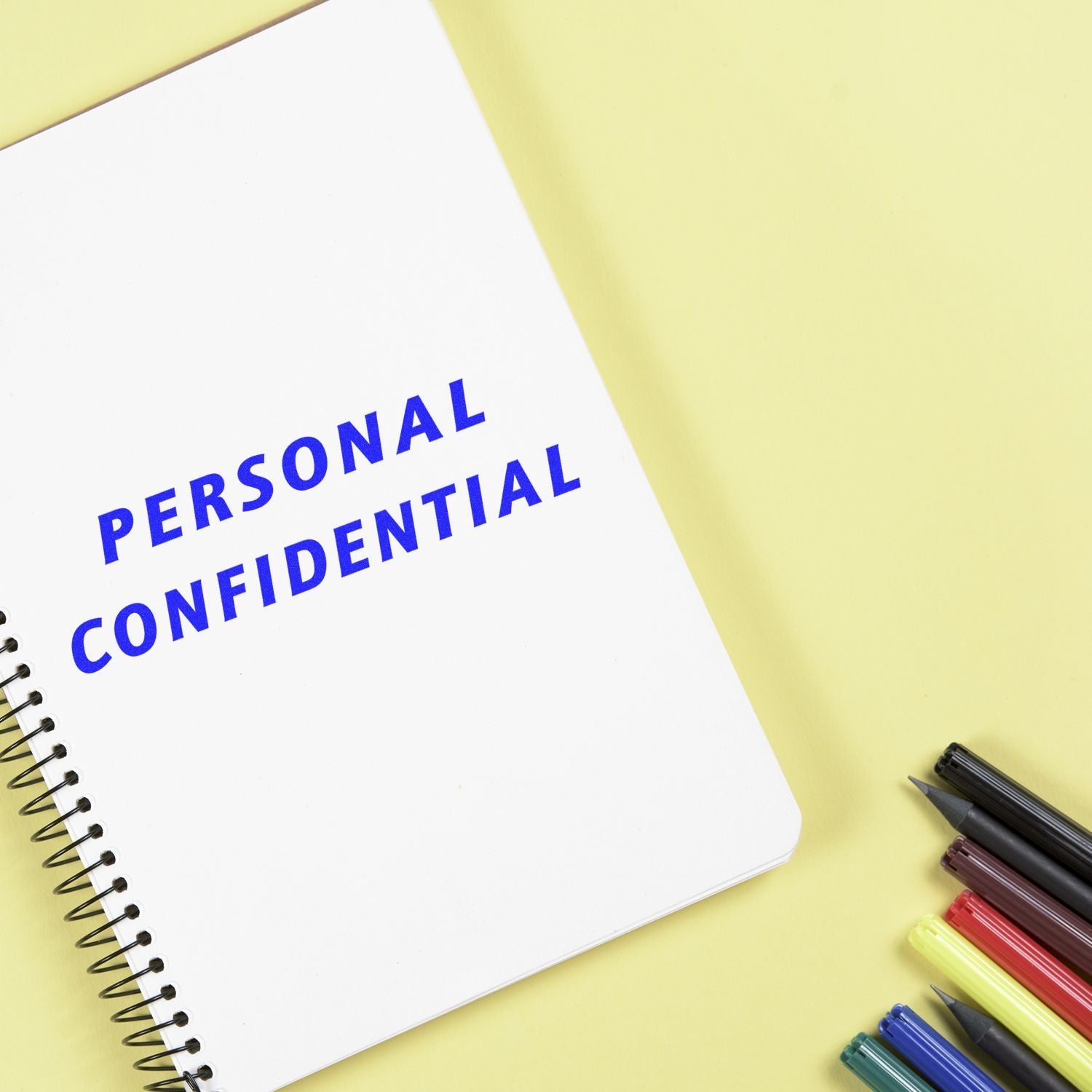 A notebook stamped with PERSONAL CONFIDENTIAL using the _Italic Personal Confidential Rubber Stamp_, next to colorful pens on a yellow surface.