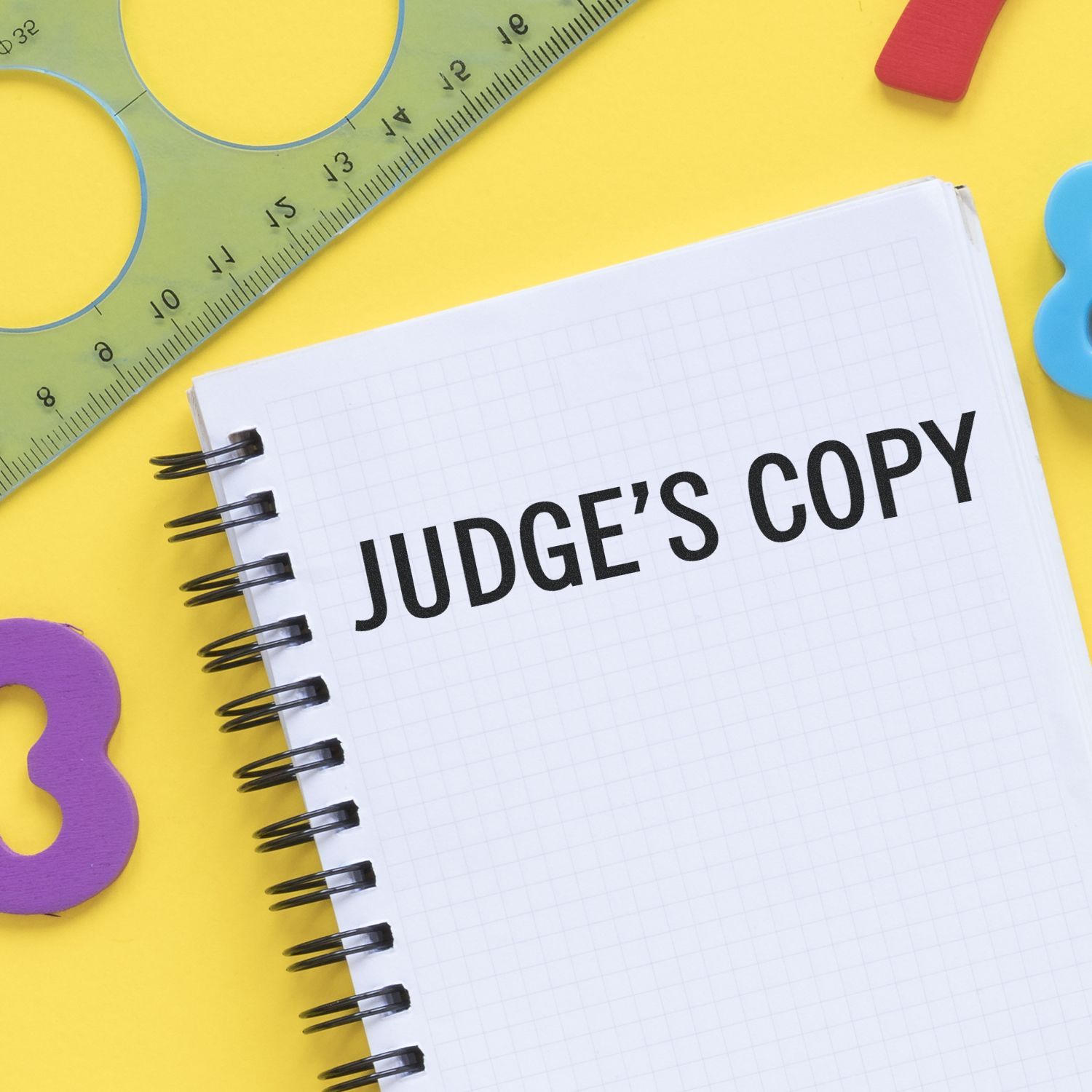 Large Pre-Inked Judge's Copy Stamp used on a spiral notebook with JUDGE'S COPY text, surrounded by colorful stationery on a yellow background.