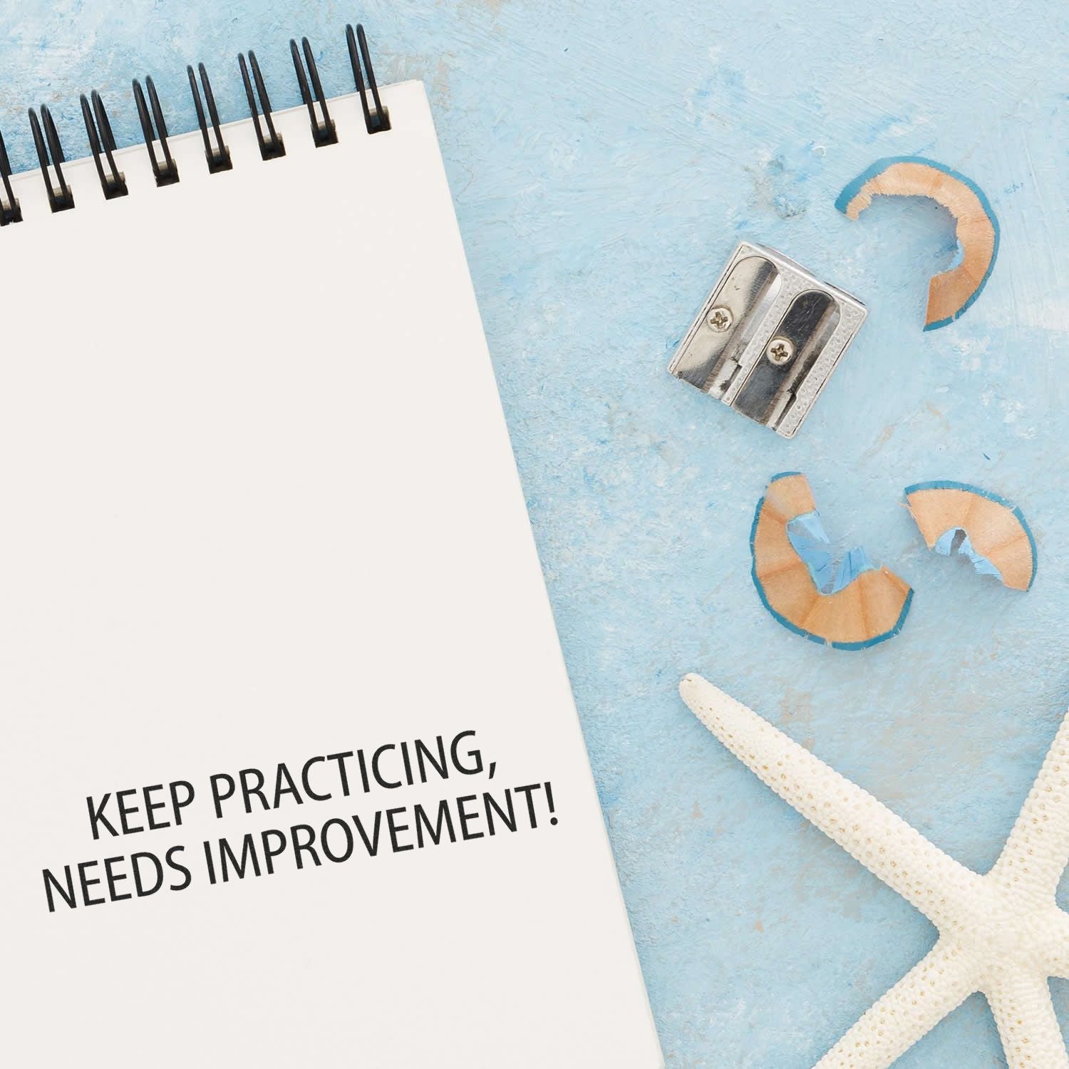 A Large Keep Practicing Needs Improvement Rubber Stamp is next to a notebook with the stamped message, a pencil sharpener, and a starfish.