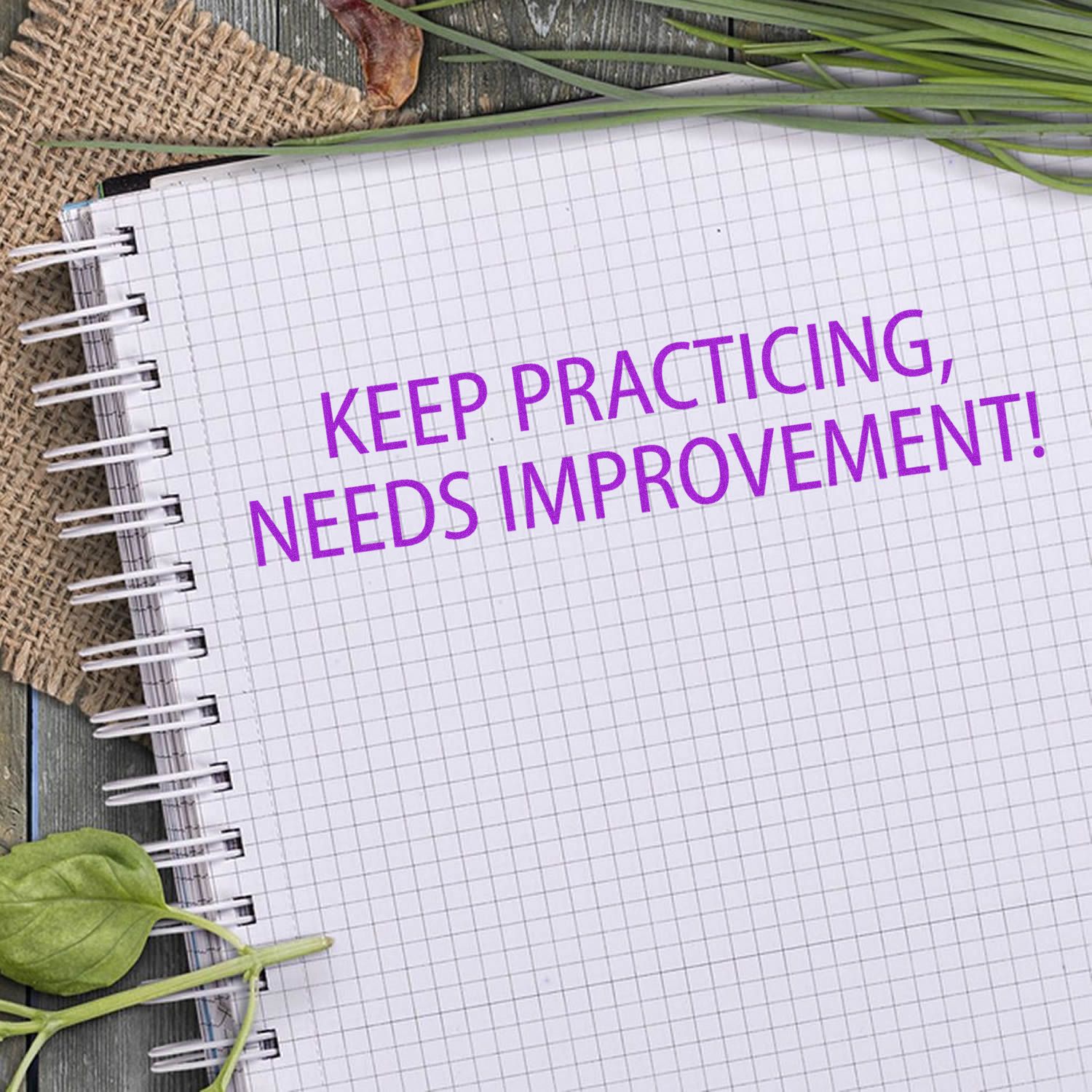 Large Keep Practicing Needs Improvement Rubber Stamp used on a spiral notebook with a grid pattern, surrounded by leaves and burlap.