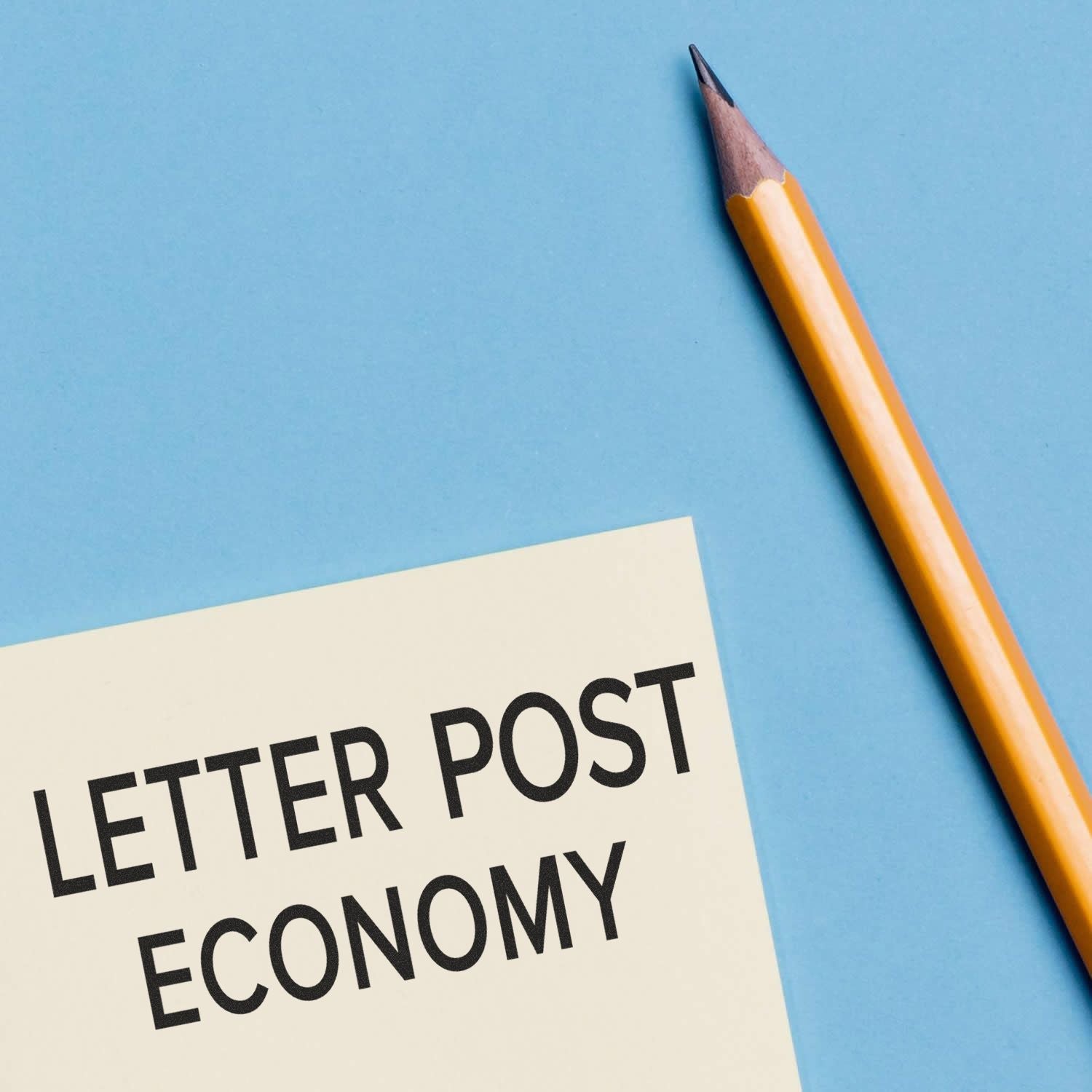 Large Pre-Inked Letter Post Economy Stamp used on a white paper next to a sharpened yellow pencil on a blue background.