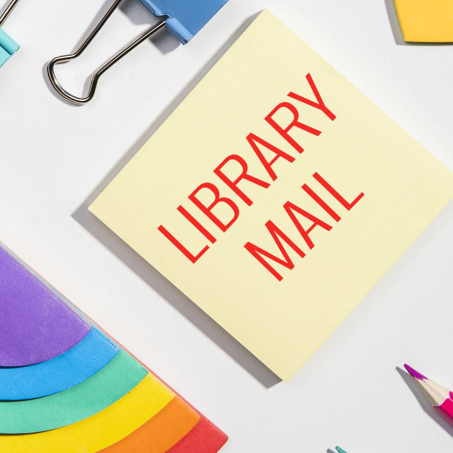 Library Mail rubber stamp on a yellow sticky note, surrounded by colorful folders, binder clips, and pencils on a white desk.