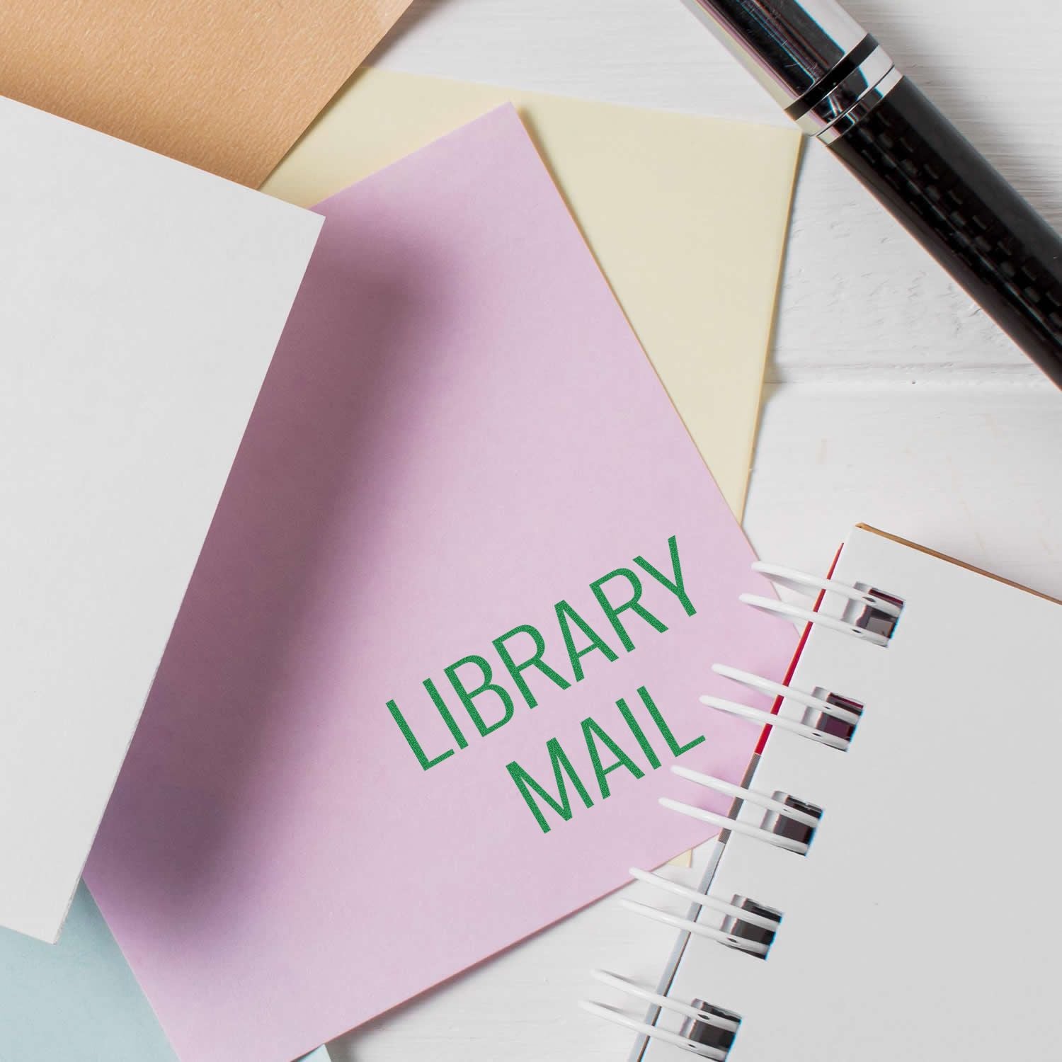 Library Mail rubber stamp in green ink on a pink envelope, surrounded by stationery and a pen on a white surface.