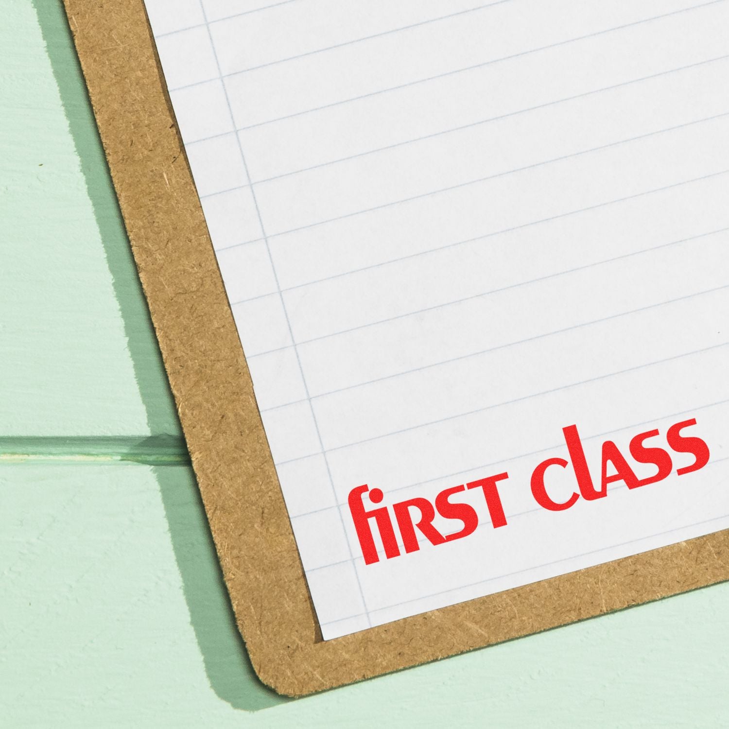 Large Pre-Inked Lower Case First Class Stamp in red ink on white lined paper clipped to a brown clipboard on a light green surface.