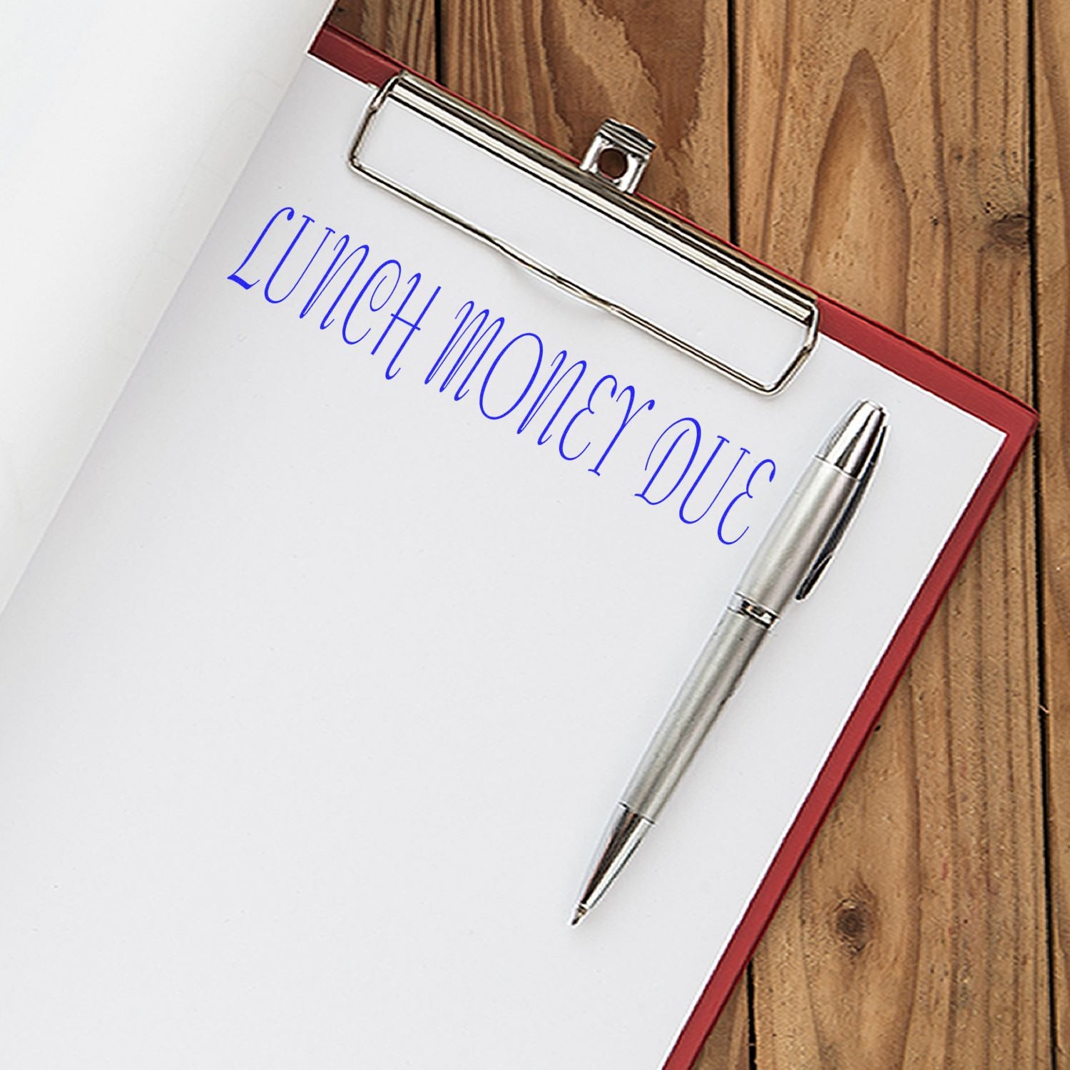 Clipboard with paper stamped LUNCH MONEY DUE using the Large Lunch Money Due Rubber Stamp, next to a silver pen on a wooden surface.