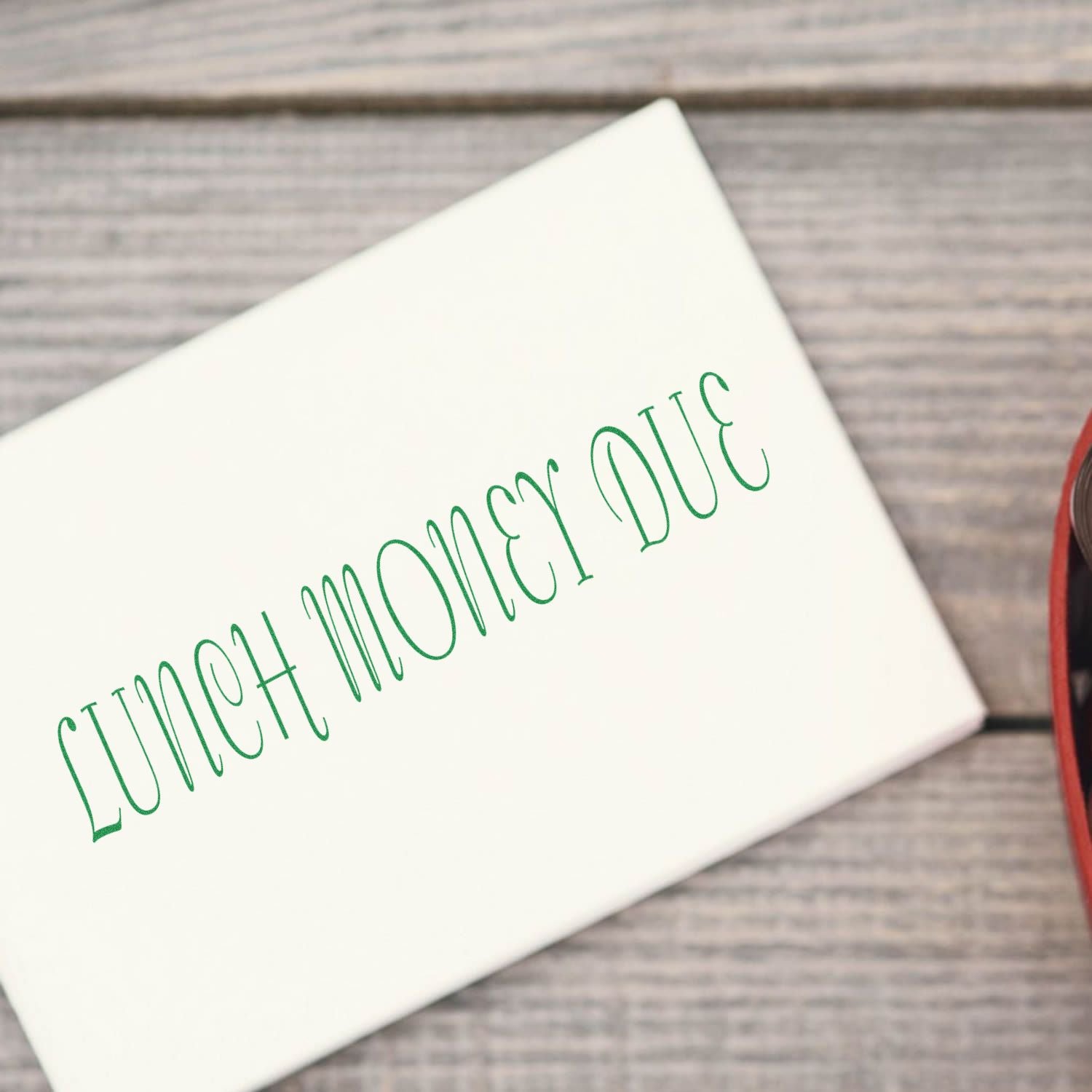 Large Self Inking Lunch Money Due Stamp in use, stamping LUNCH MONEY DUE in green on a white envelope, placed on a wooden surface.