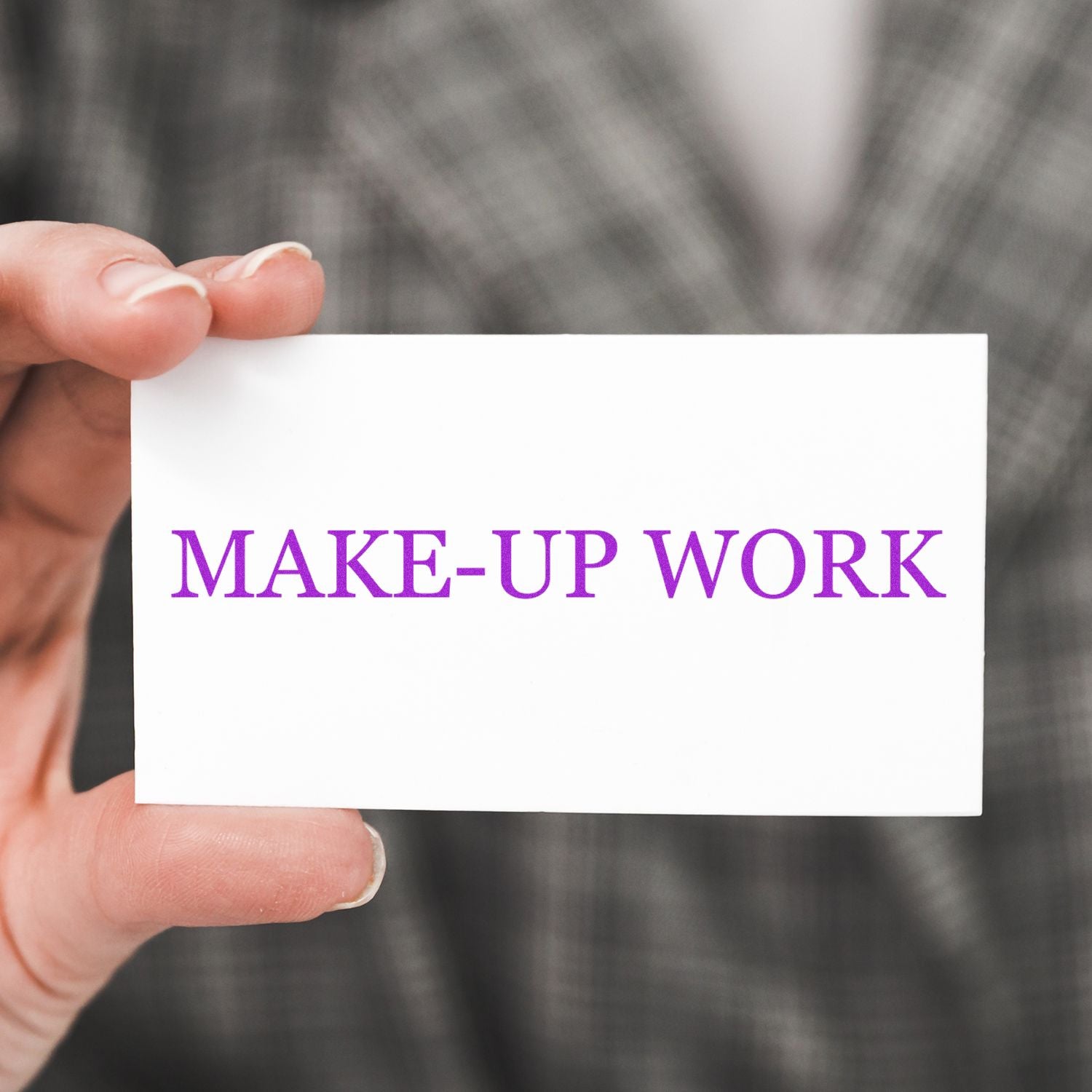 Hand holding a white card stamped with MAKE-UP WORK in purple using the Large Make Up Work Rubber Stamp, against a blurred plaid background.