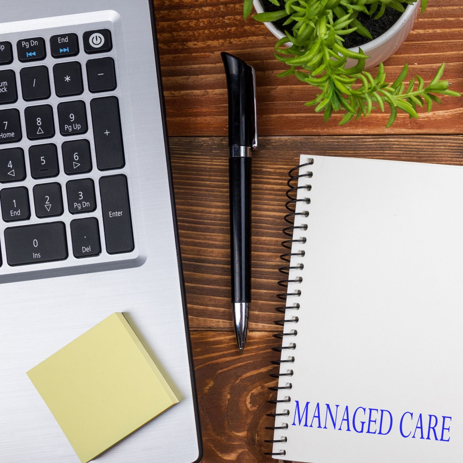 Large Self Inking Managed Care Stamp in use on a spiral notebook, placed on a wooden desk with a laptop, pen, sticky notes, and a plant.