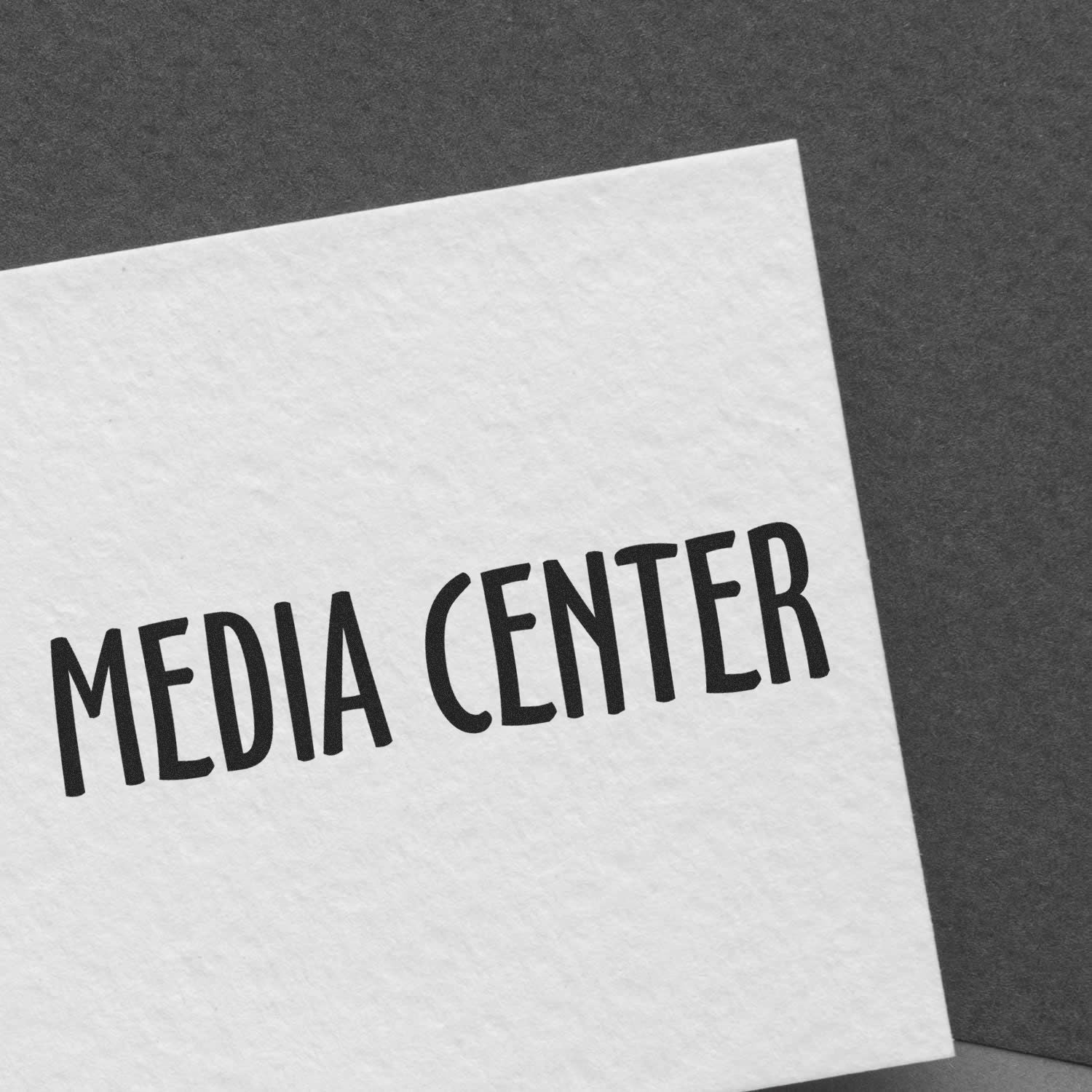 A white card stamped with MEDIA CENTER using the Large Pre-Inked Media Center Stamp, placed on a dark gray surface.