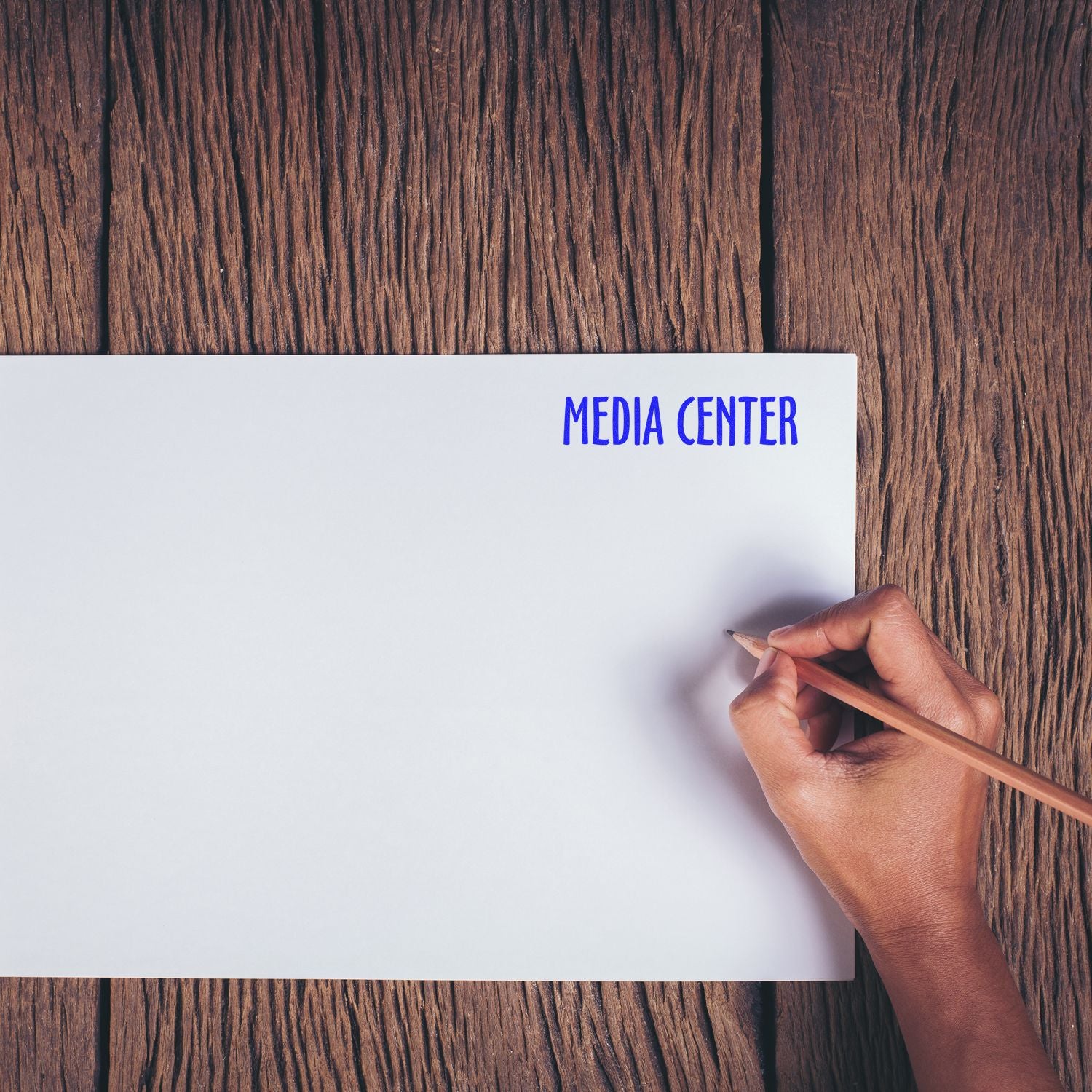 Hand holding pencil near paper stamped with Media Center in blue ink on a wooden surface.