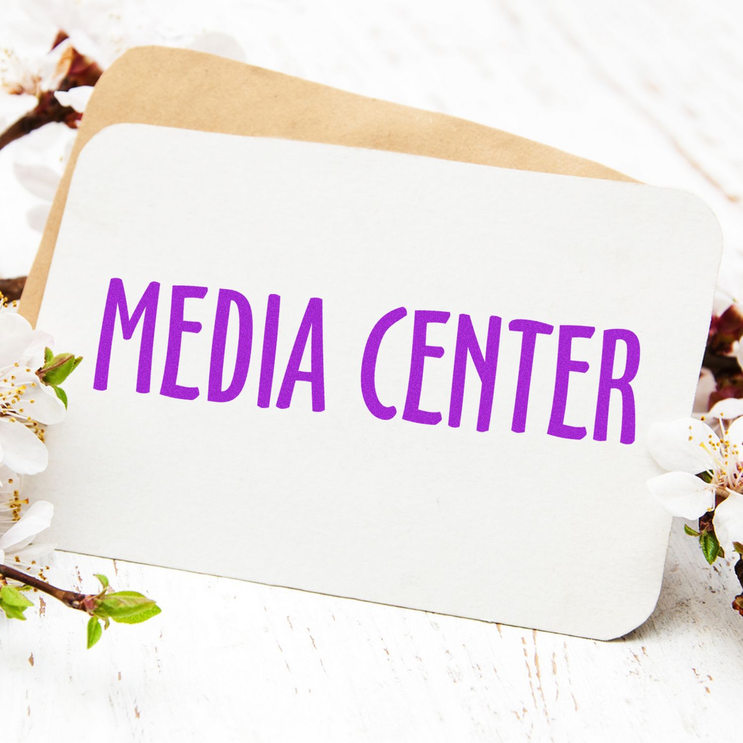 A card with Media Center stamped in purple ink, surrounded by white flowers and a brown envelope in the background.