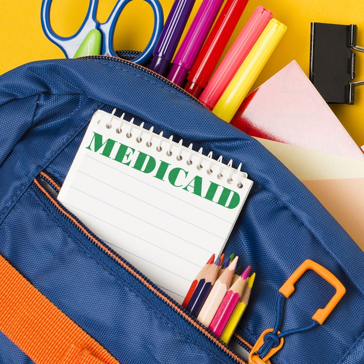 A Self Inking Medicaid Stamp is shown in use on a notepad, placed inside a blue backpack with colorful stationery items.