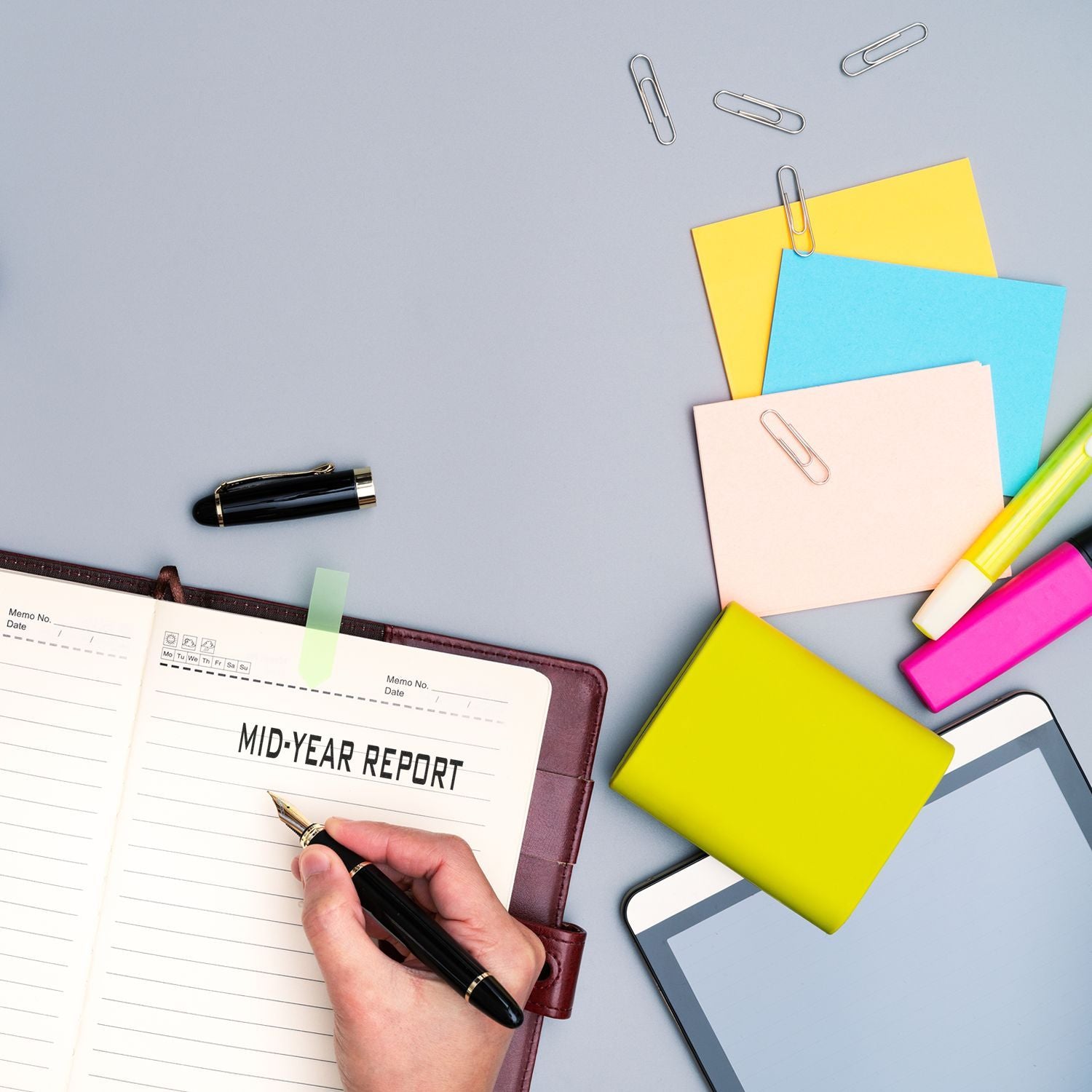 Person using the Large Self Inking Mid Year Report Stamp on a notebook, with colorful sticky notes, pens, and a tablet on a desk.