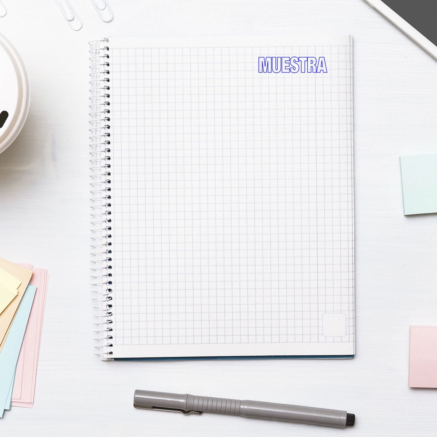 Large Muestra Rubber Stamp used on a spiral notebook with grid paper, surrounded by a pen, sticky notes, and a smartphone on a white desk.