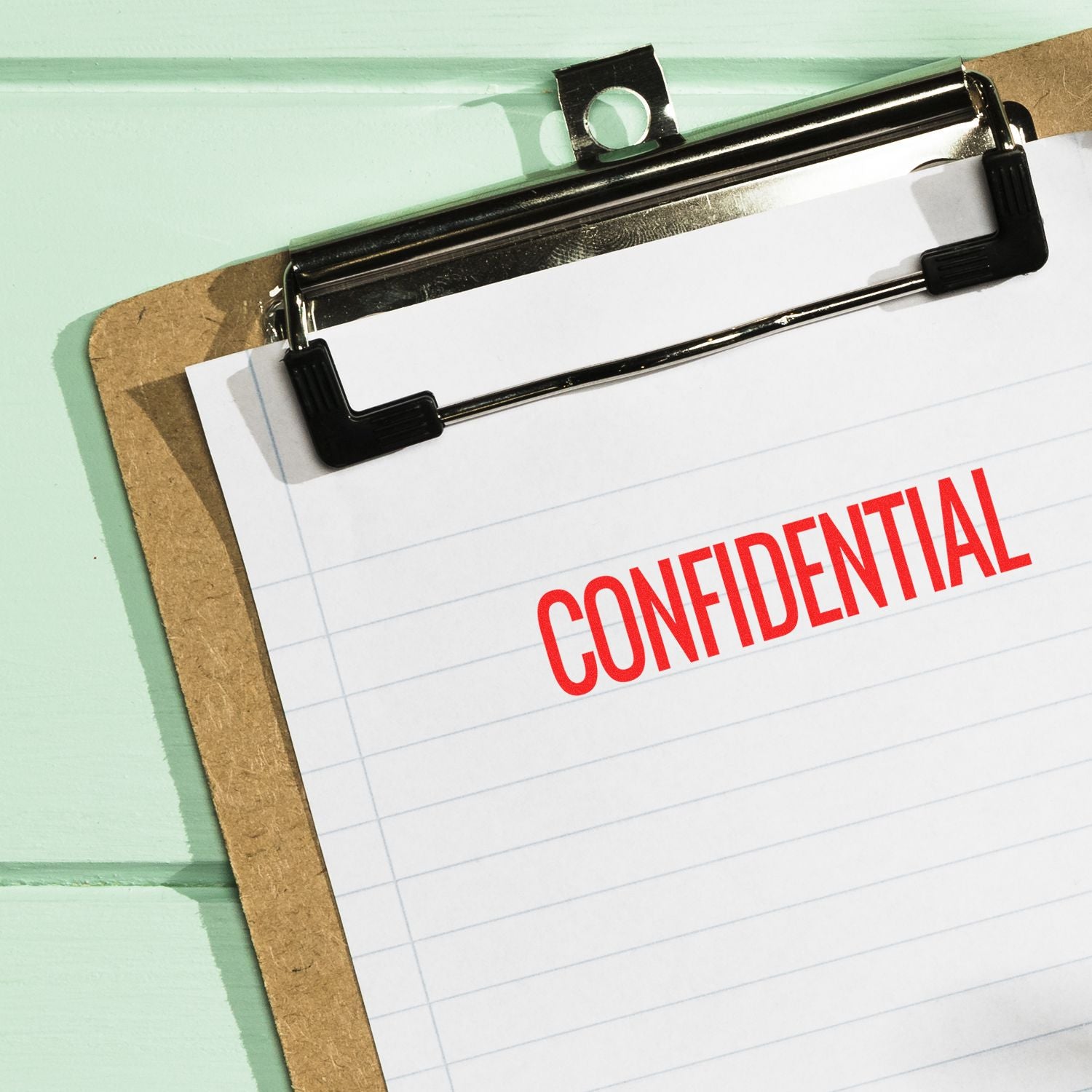 Clipboard with lined paper stamped CONFIDENTIAL in red using the Large Self Inking Narrow Confidential Stamp, placed on a mint green background.