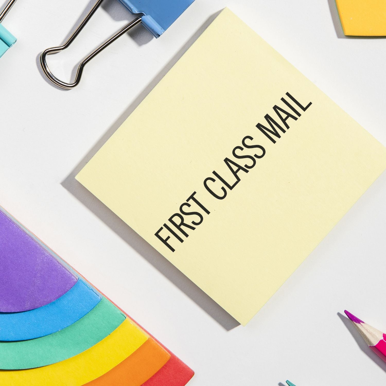 Large Pre-Inked Narrow First Class Mail Stamp used on a yellow sticky note, surrounded by colorful office supplies on a white desk.