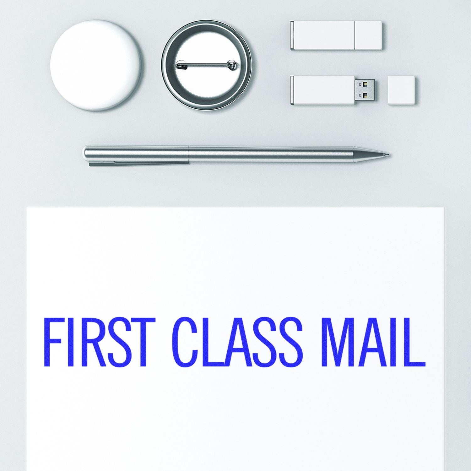 Large Pre-Inked Narrow First Class Mail Stamp used on white paper, surrounded by a pen, USB drives, and round objects on a white surface.