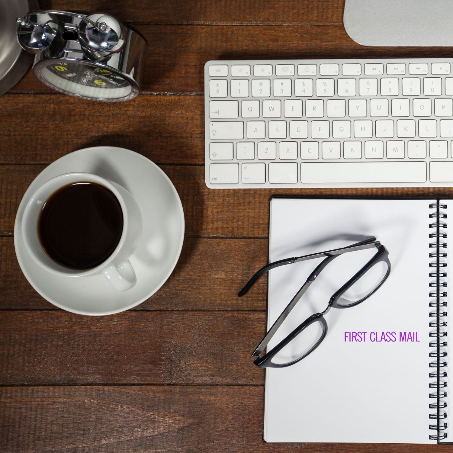 A desk with a keyboard, coffee, glasses, and a notebook stamped with FIRST CLASS MAIL using the Large Self Inking Narrow First Class Mail Stamp.