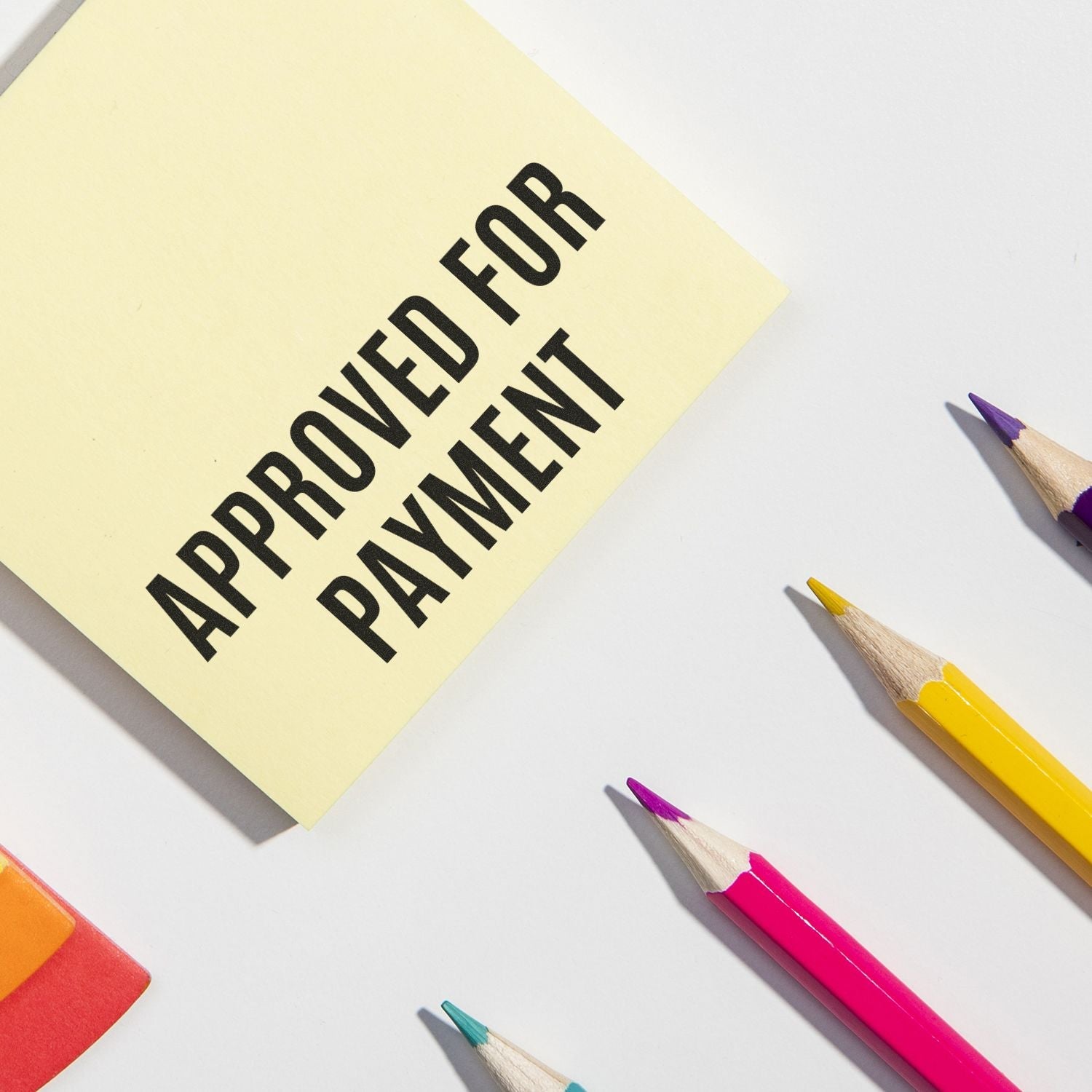 Self Inking Narrow Font Approved for Payment Stamp on a yellow sticky note, surrounded by colorful pencils and an eraser on a white background.