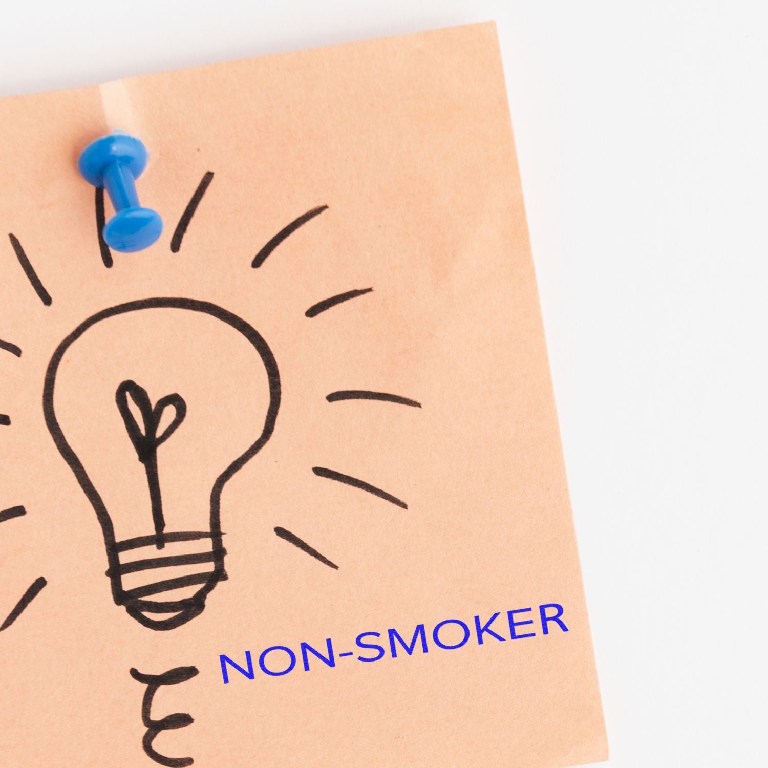 A Large Self Inking Narrow Font Non-Smoker Stamp marks NON-SMOKER in blue ink on a paper with a lightbulb drawing, pinned with a blue pushpin.