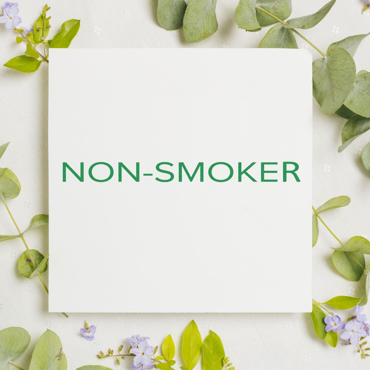 Slim Pre-Inked Narrow Font Non-Smoker Stamp in green ink on white paper, surrounded by green leaves and small purple flowers.