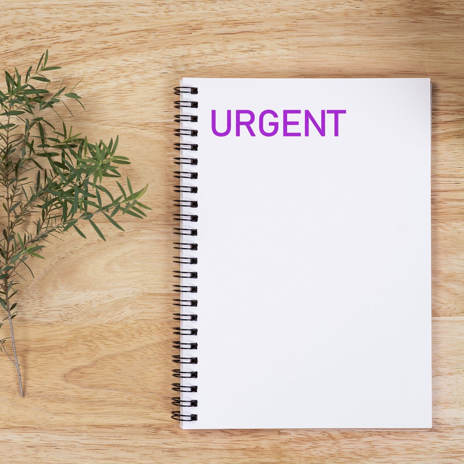 Large Narrow Font Urgent Rubber Stamp in purple ink on a white spiral notebook, placed on a wooden surface with a small plant beside it.