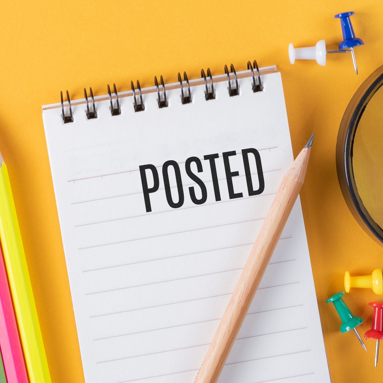 Large Self Inking Narrow Posted Stamp marking POSTED on a spiral notepad, surrounded by colorful stationery on a yellow background.