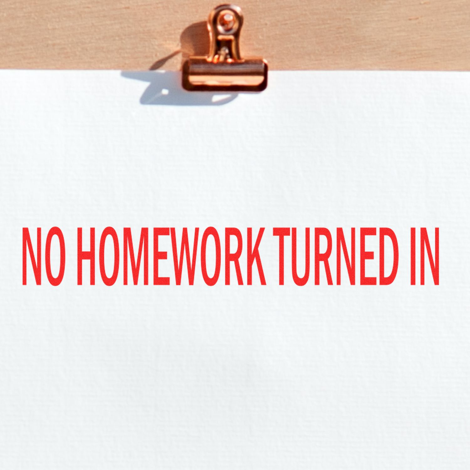 A paper clipped to a wooden board with a red NO HOMEWORK TURNED IN stamp impression using the Large No Homework Turned In Rubber Stamp.