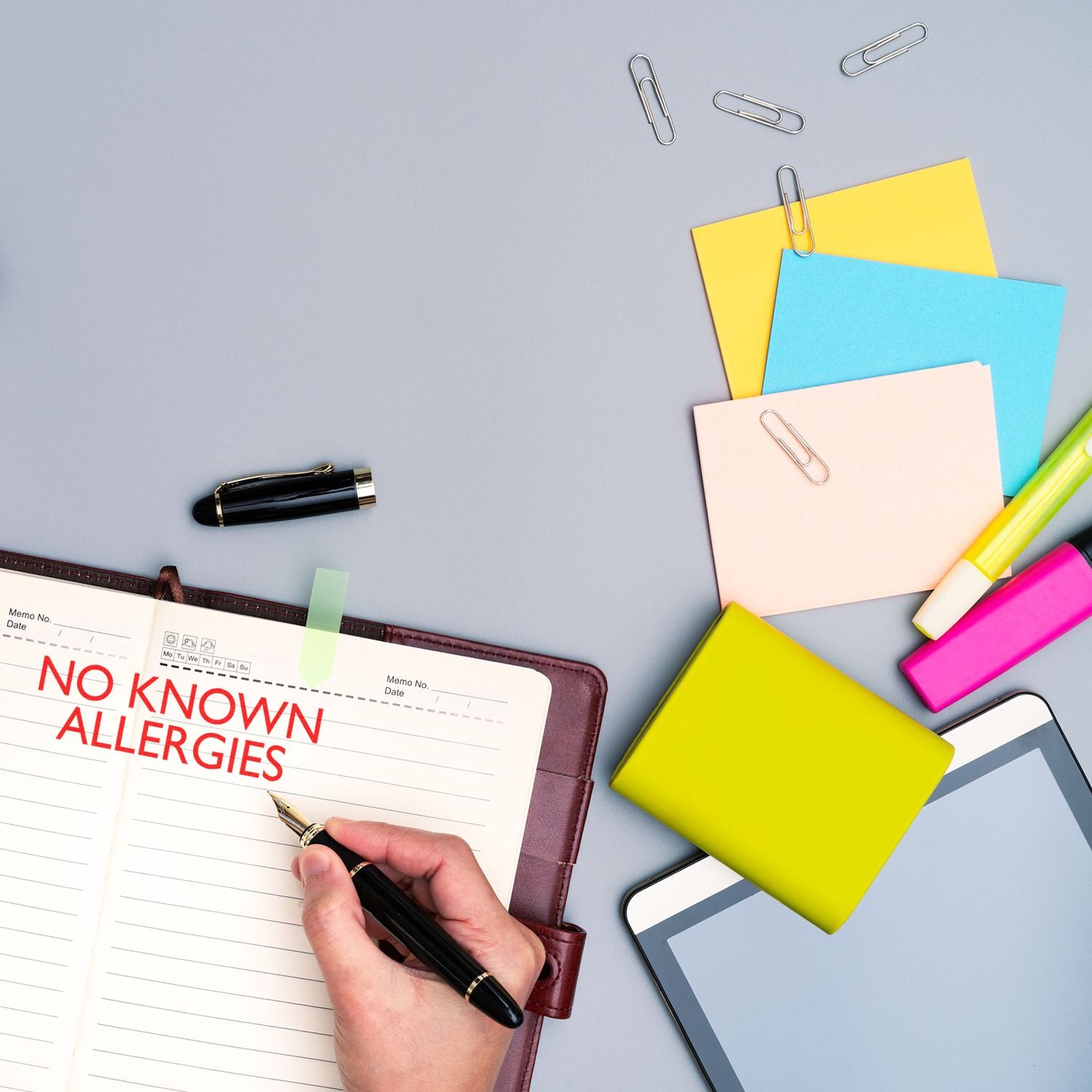 Person using a Slim Pre-Inked No Known Allergies Stamp on a notebook, surrounded by colorful stationery and a tablet.