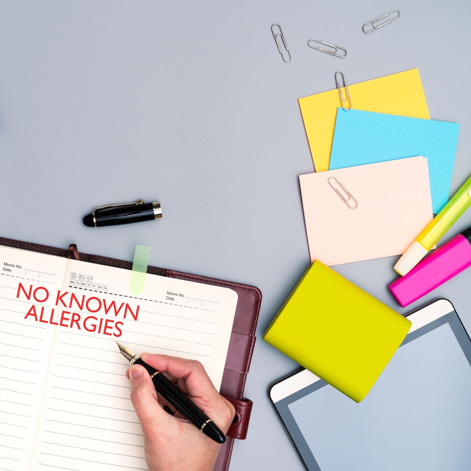 A hand using the Self Inking No Known Allergies Stamp on a notebook, surrounded by colorful sticky notes, paper clips, and office supplies.