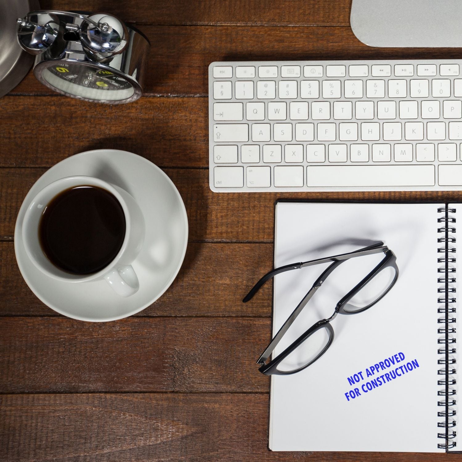 Self Inking Not Approved For Construction Stamp used on a notebook, placed on a wooden desk with a keyboard, glasses, coffee, and clock.