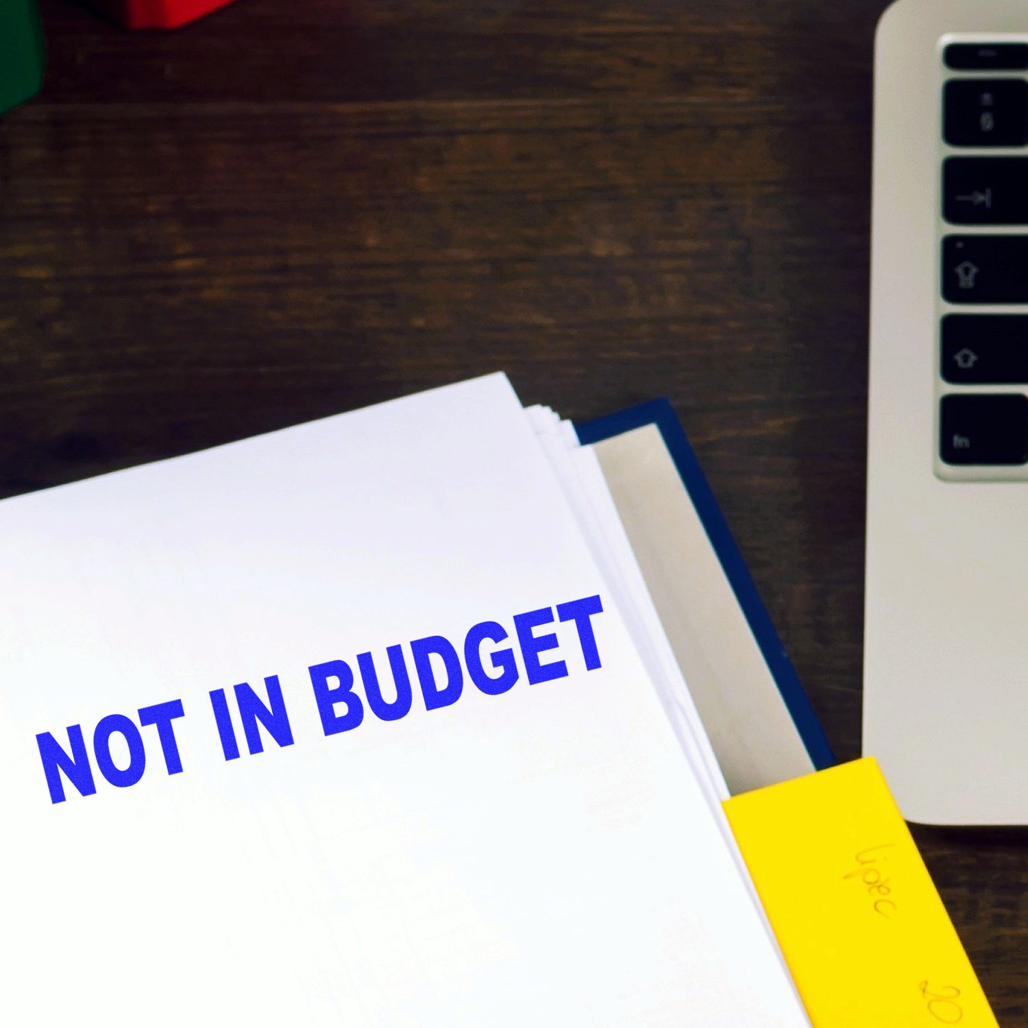 Large Not In Budget Rubber Stamp marking a document on a wooden desk next to a laptop and a yellow sticky note.