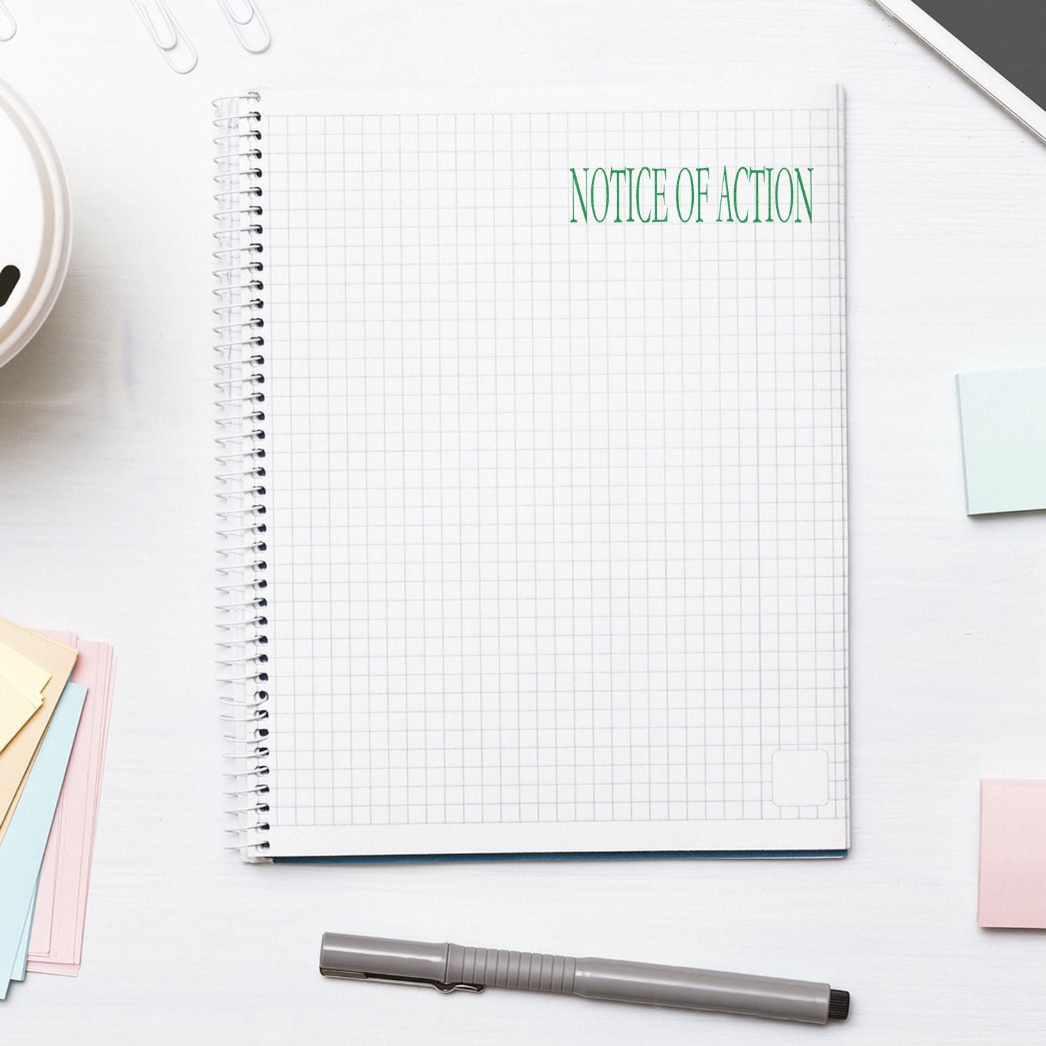 Large Self Inking Notice Of Action Stamp in green ink on a spiral notebook, surrounded by office supplies on a white desk.