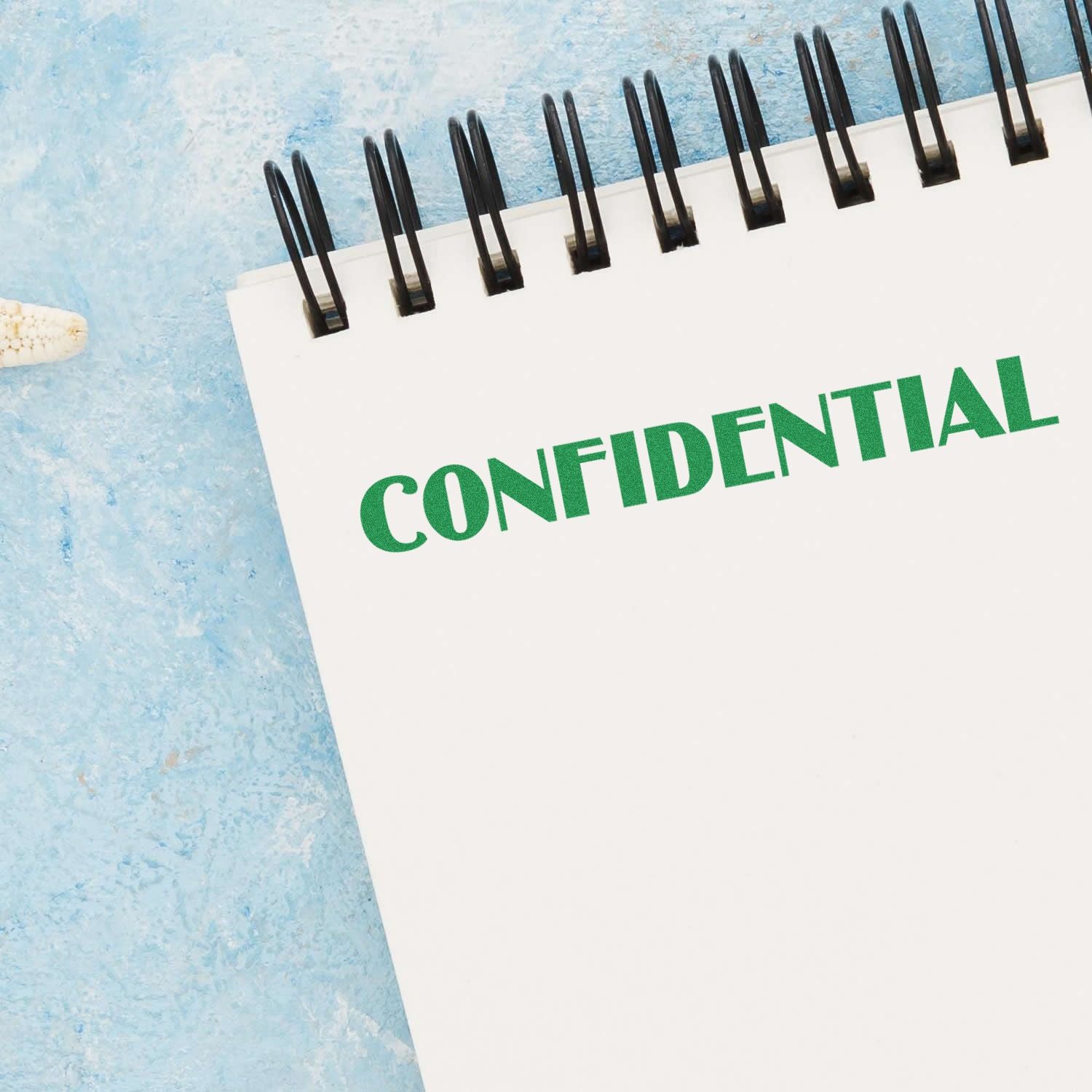 Large Optima Confidential Rubber Stamp marking CONFIDENTIAL in green on a white spiral notebook against a blue textured background.