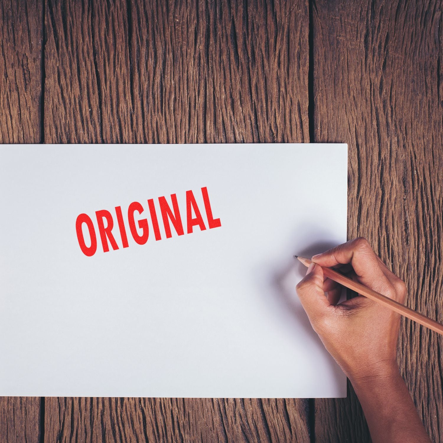 Hand holding a pencil next to a paper stamped with a red Original rubber stamp on a wooden surface.