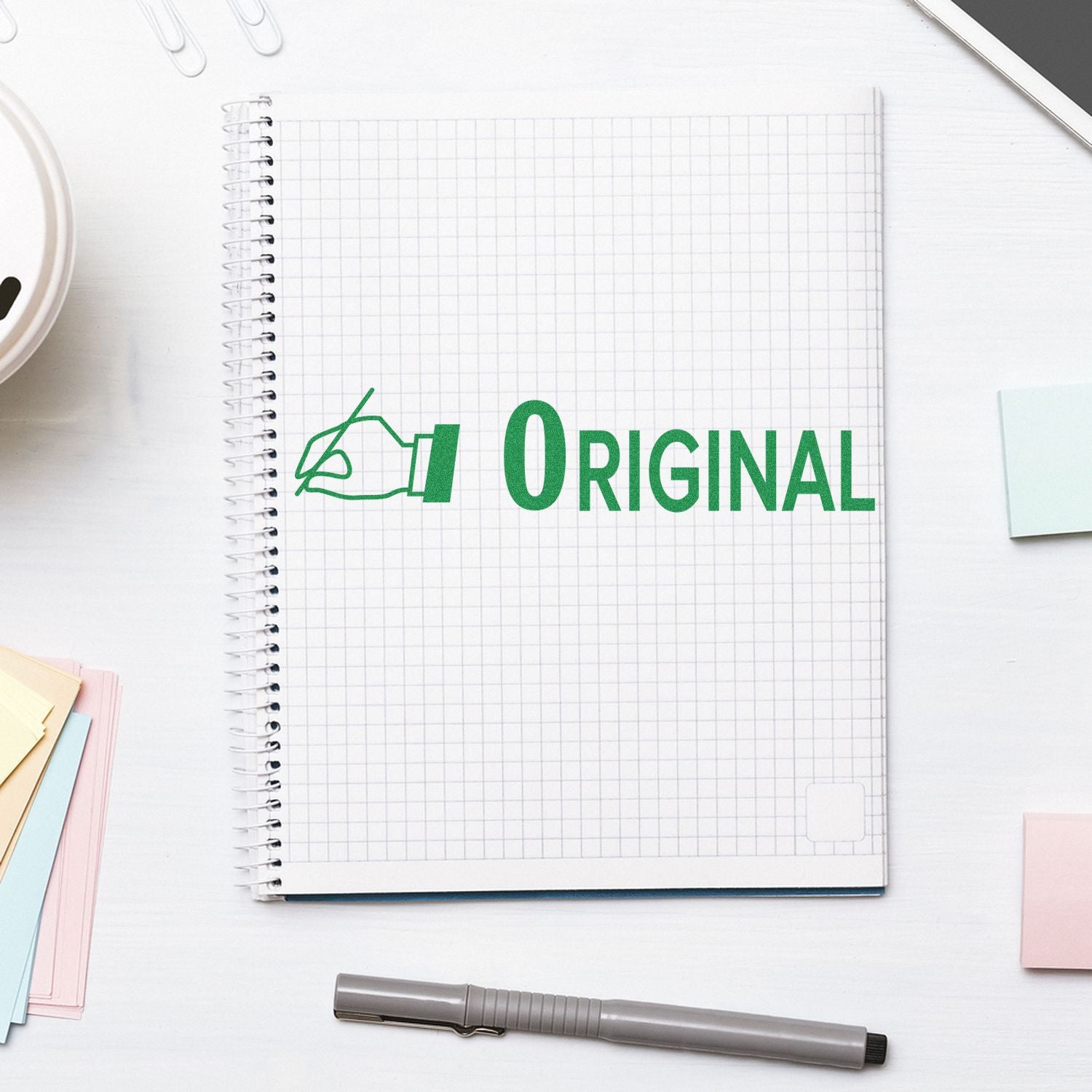 A notebook with a grid pattern displays a green Original stamp from the Large Pre-Inked Original with Hand Stamp, surrounded by office supplies.