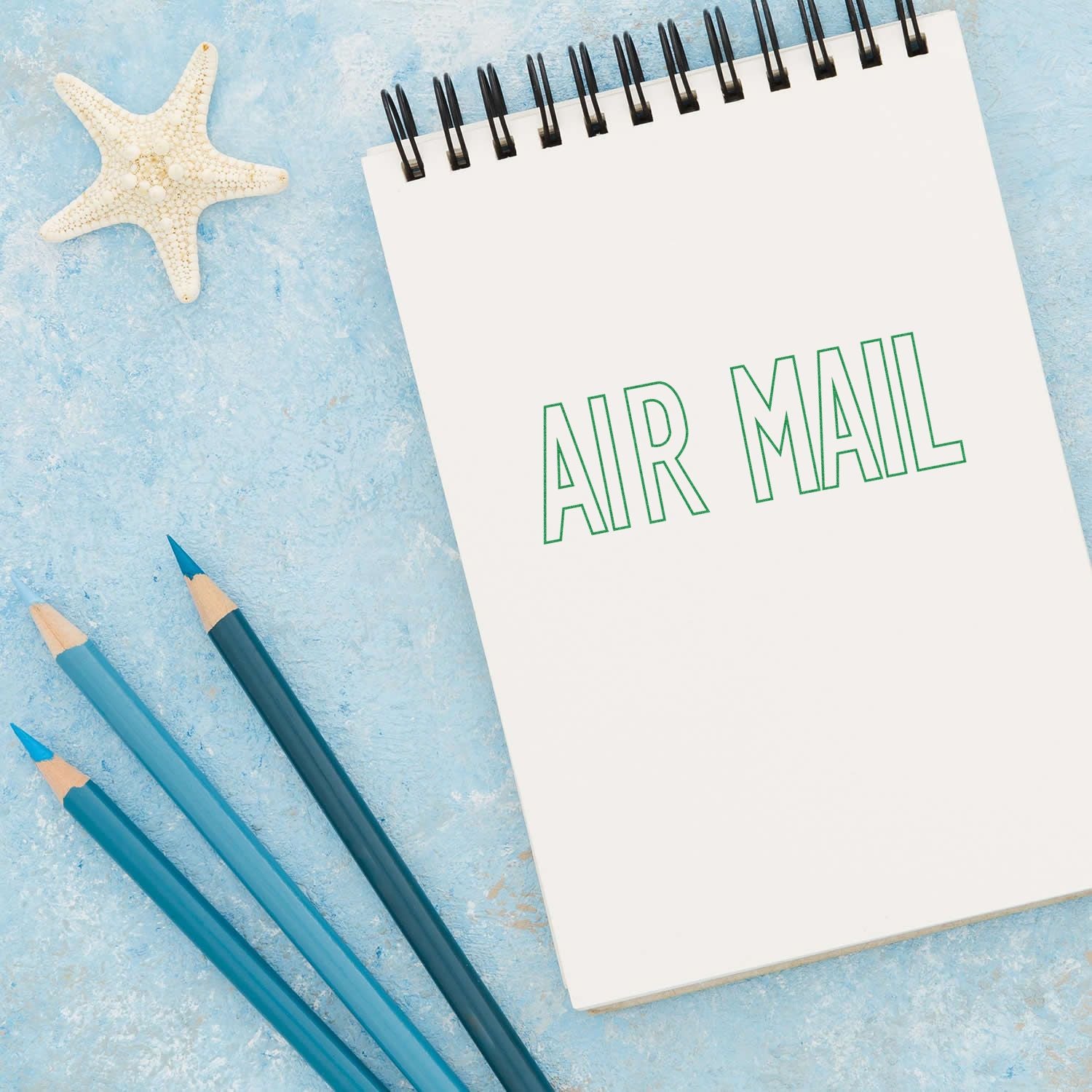 Large Self Inking Outline Air Mail Stamp used on a white notepad with AIR MAIL text, next to blue pencils and a starfish on a blue background.