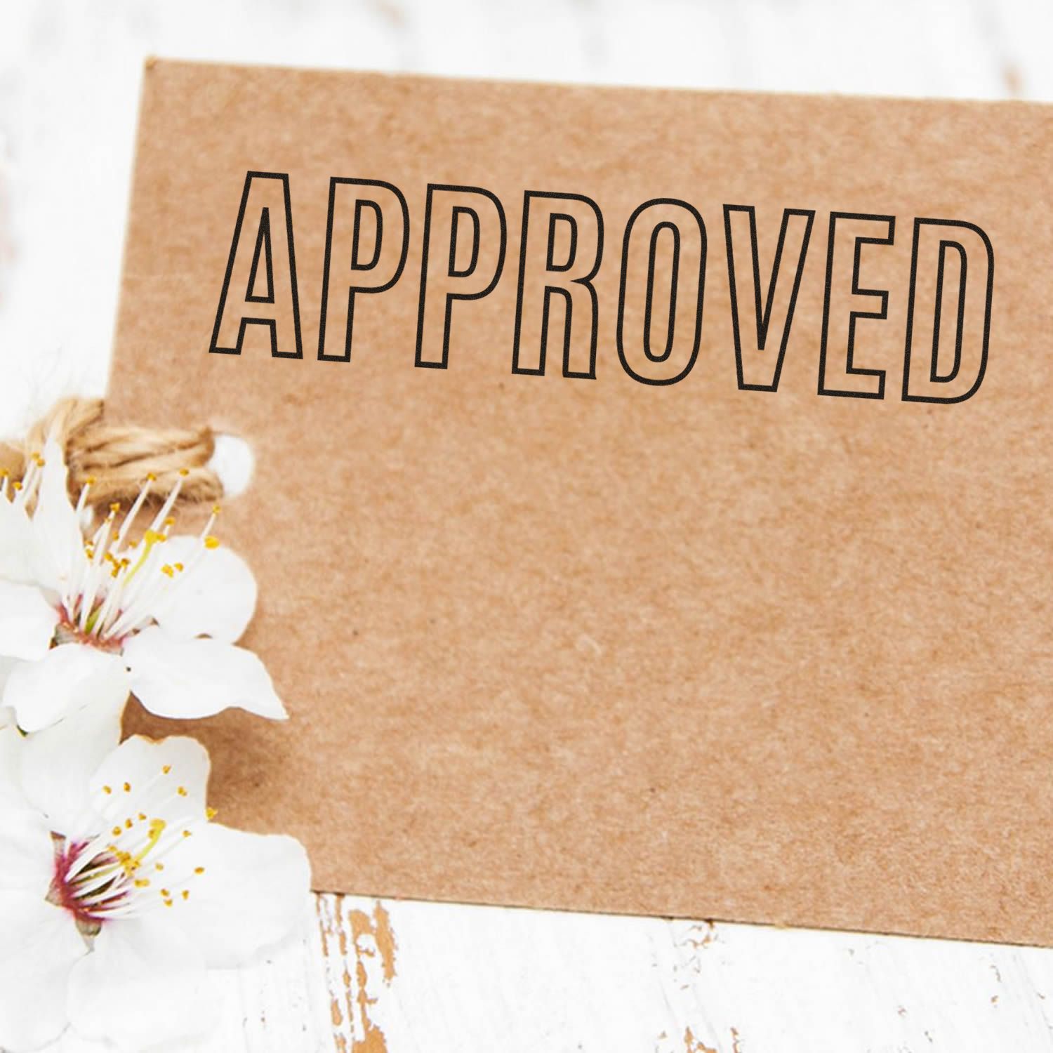 A brown card stamped with APPROVED using the Large Pre-Inked Outline Approved Stamp, placed next to white flowers on a white wooden surface.