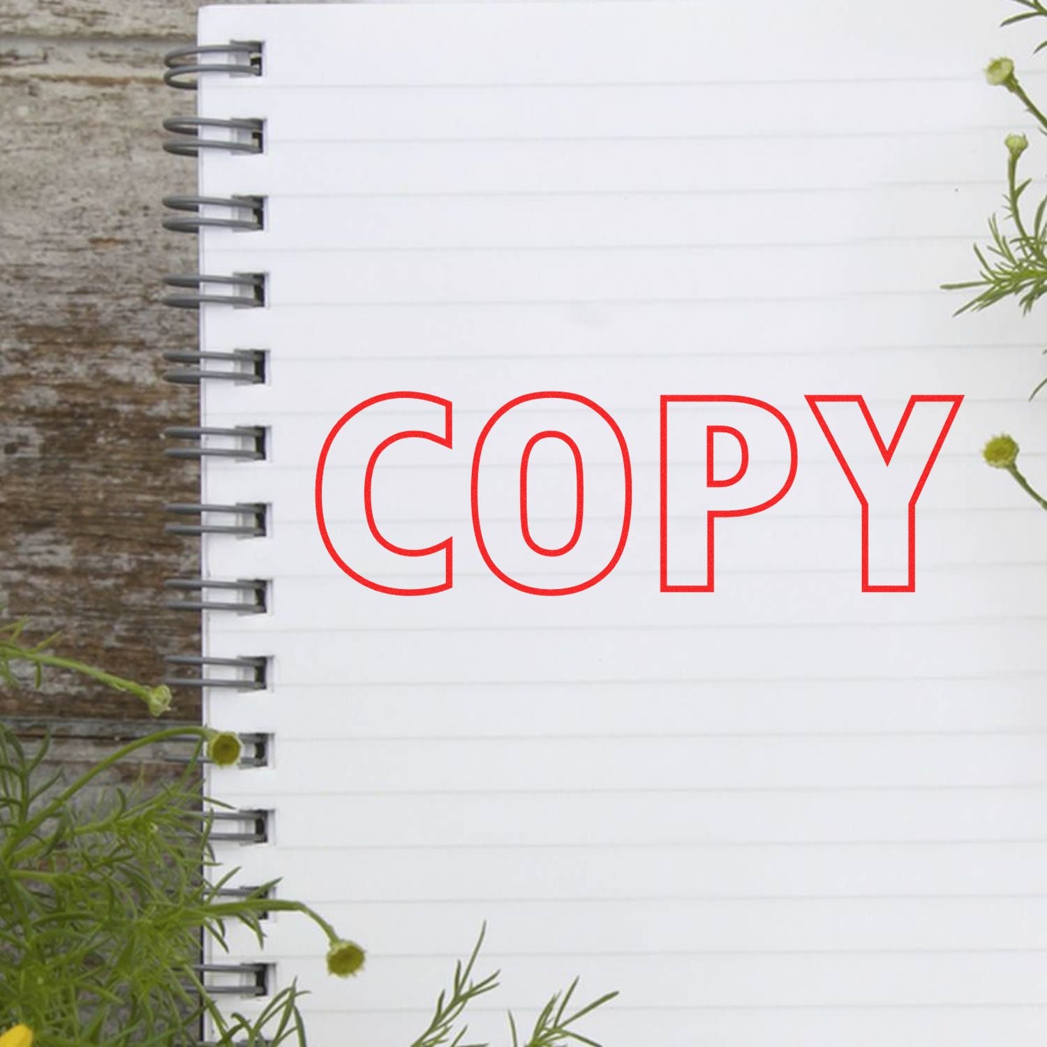 Copy Outline Text Rubber Stamp in red ink on a white spiral notebook with lined pages, surrounded by green plants.