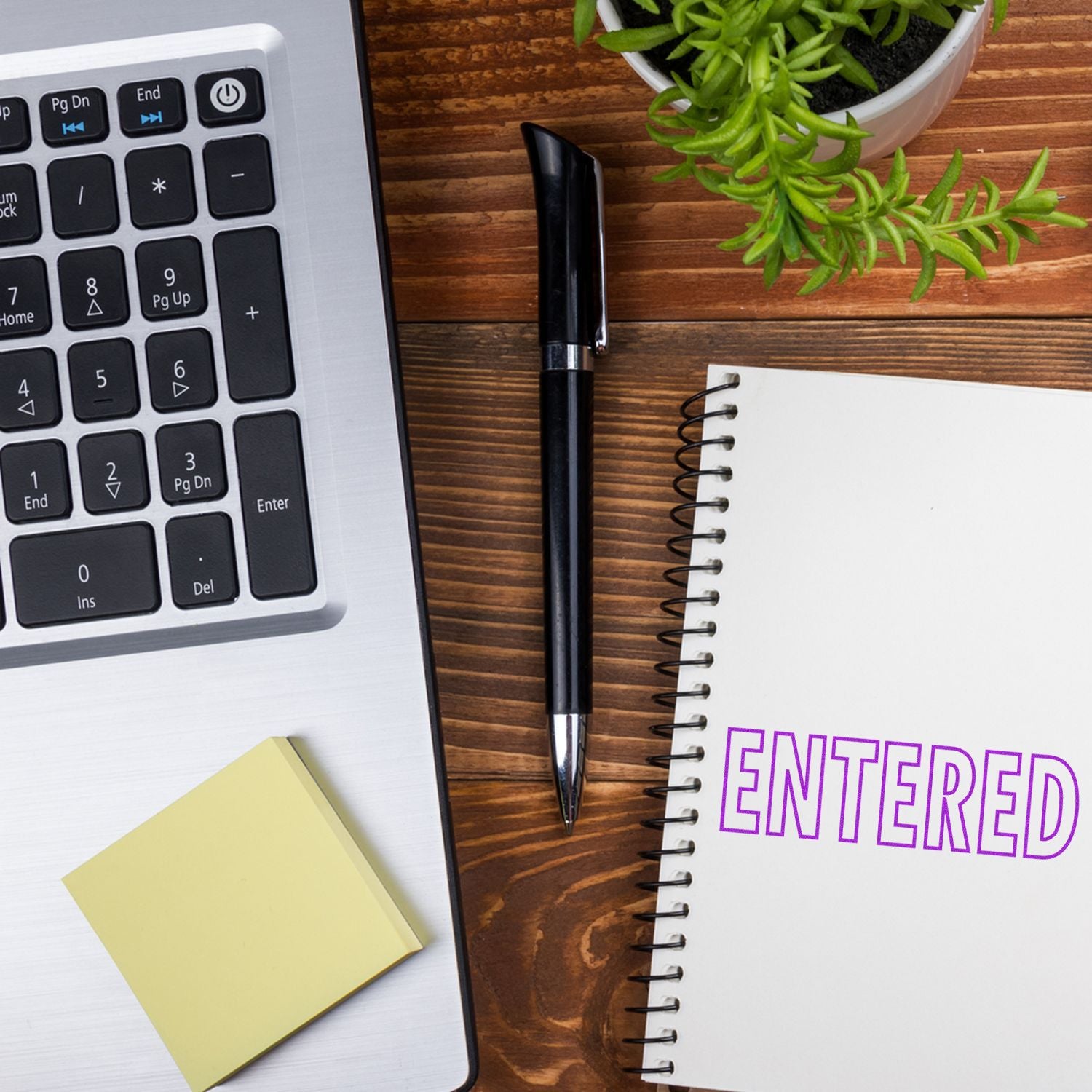 Desk with a laptop, sticky notes, pen, plant, and notebook with 'Entered' stamped in purple using an Outline Entered Rubber Stamp.