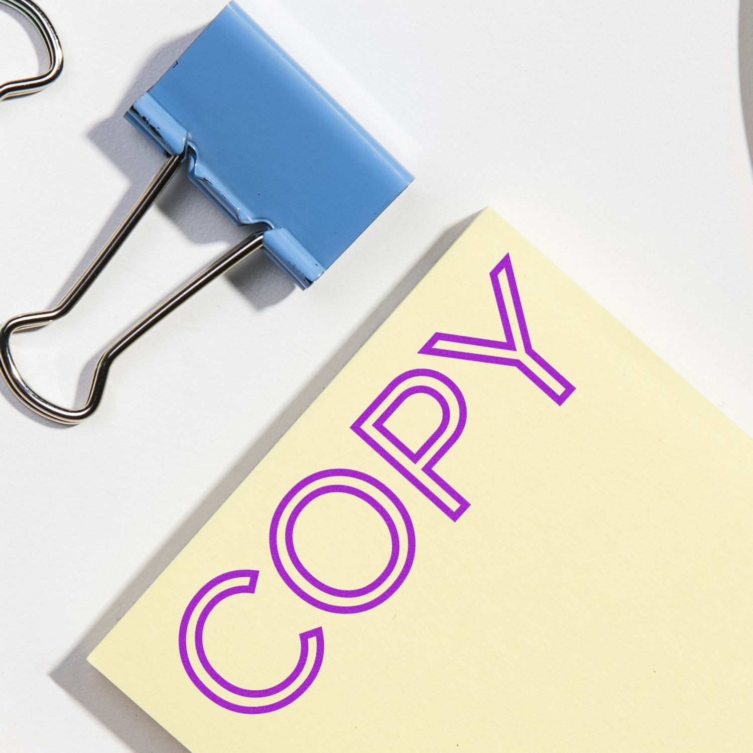 Self Inking Outline Font Copy Stamp marking COPY in purple on a yellow paper, next to a blue binder clip on a white surface.