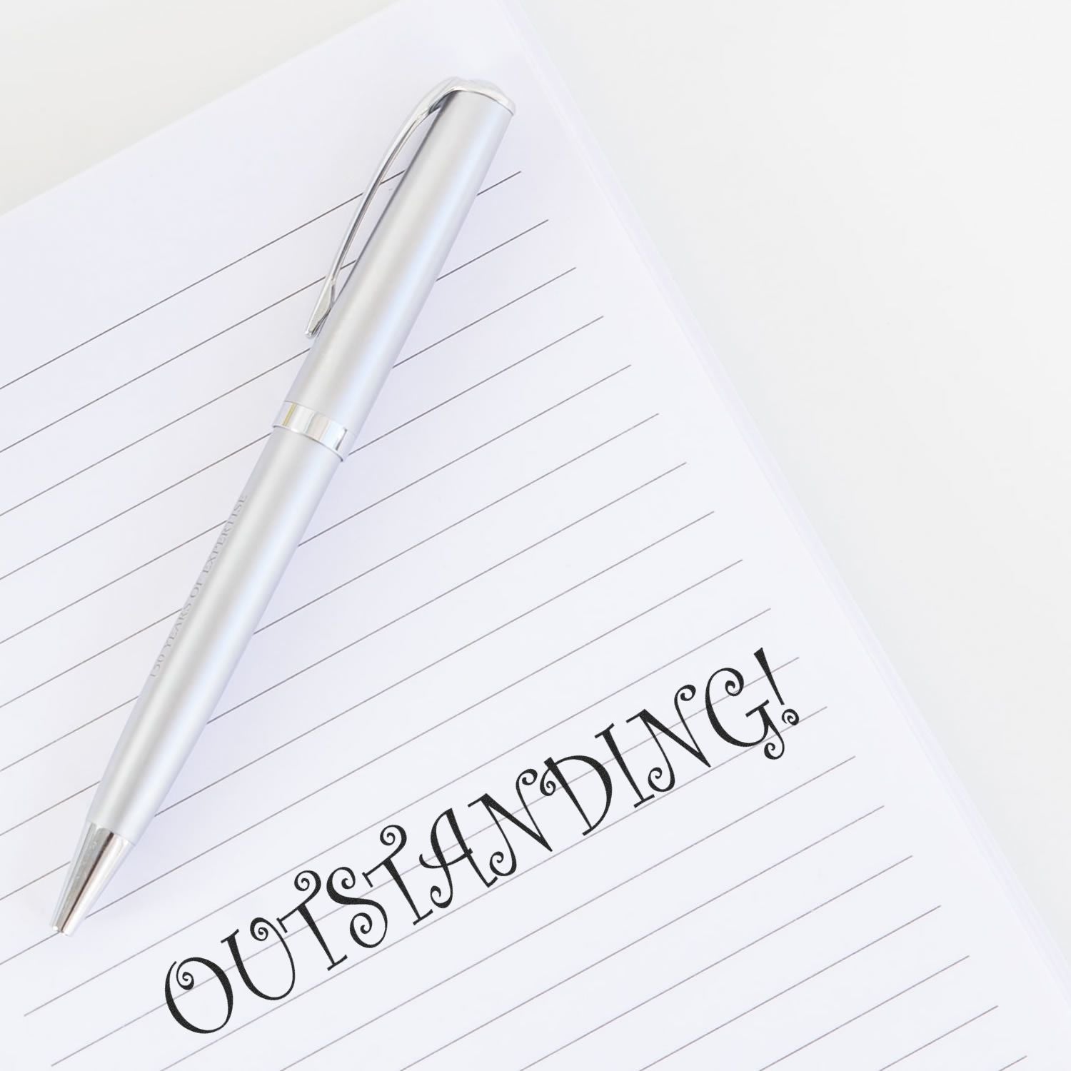 A silver pen rests on a lined notebook with the word OUTSTANDING! stamped in bold letters using the Large Outstanding Rubber Stamp.