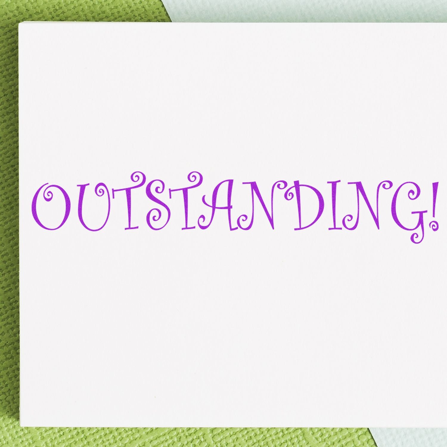 A white card with the word 'OUTSTANDING!' stamped in purple using an Outstanding Rubber Stamp, placed on a green textured surface.