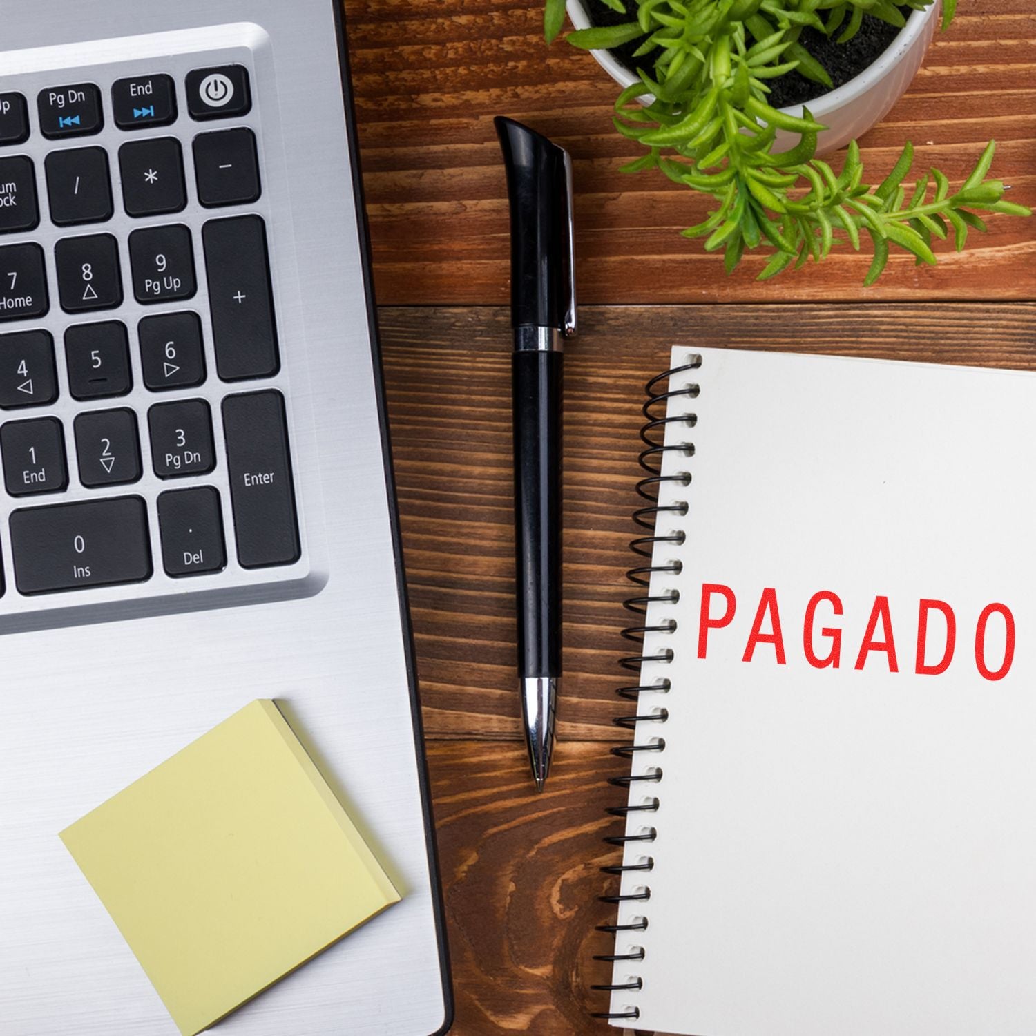 Large Pre-Inked Pagado Stamp used on a notebook, placed on a wooden desk with a laptop, pen, sticky notes, and a potted plant.