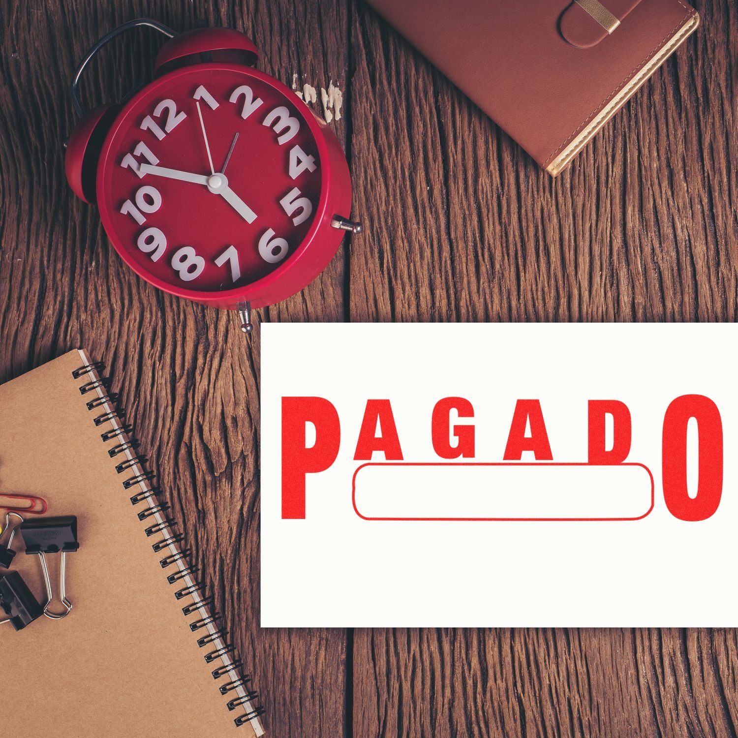 Large Pagado with Box Rubber Stamp in use on a white paper, placed on a wooden desk with a red clock, notebook, and office supplies.